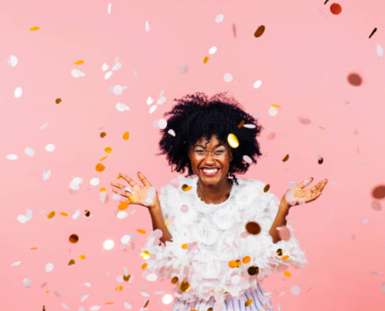 Joyful woman with curly hair celebrating amidst pink background with colorful confetti falling around her.