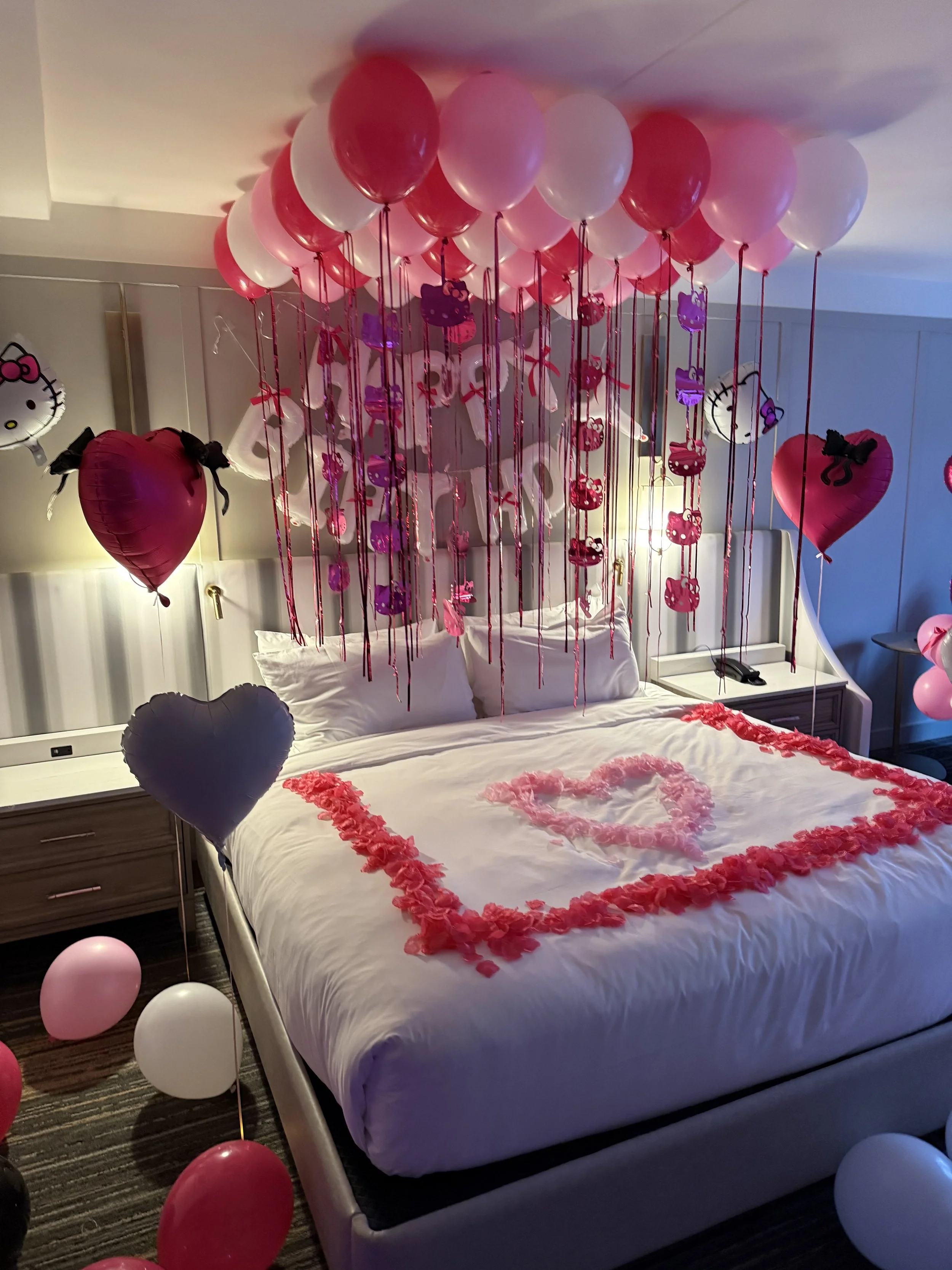 A decorated hotel room heart-themed decorations with pink, red, black, and white balloons, including hanging balloons with Hello Kitty and other character cutouts. The bed has a heart outline made of pink rose petals on the white bedding.