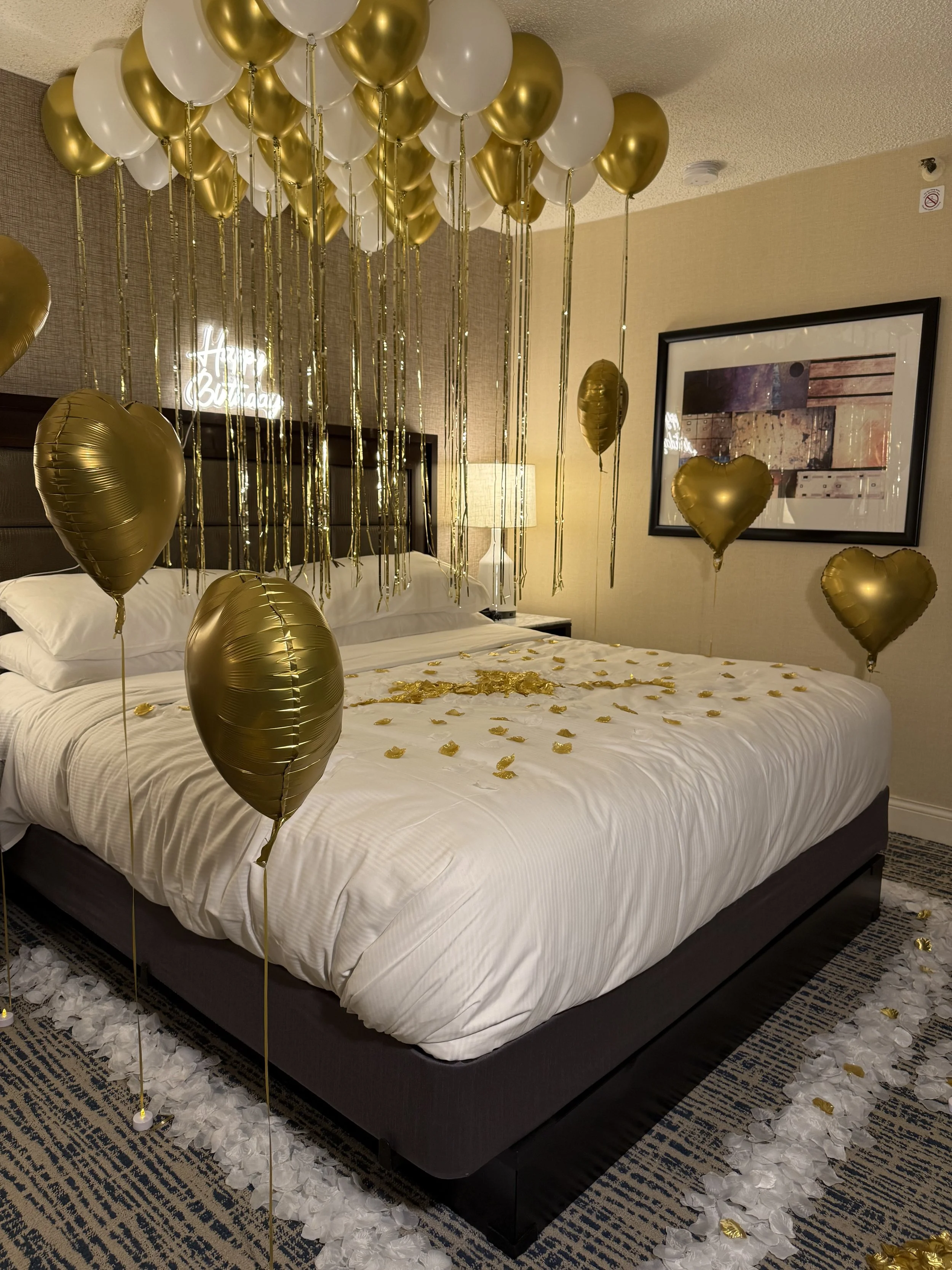 A hotel room decorated for a celebration with gold and white balloons hanging from the ceiling and surrounding the bed. Rose petals and gold confetti are scattered on the bed, and a ring of rose petals with gold accents decorates the floor around the bed.