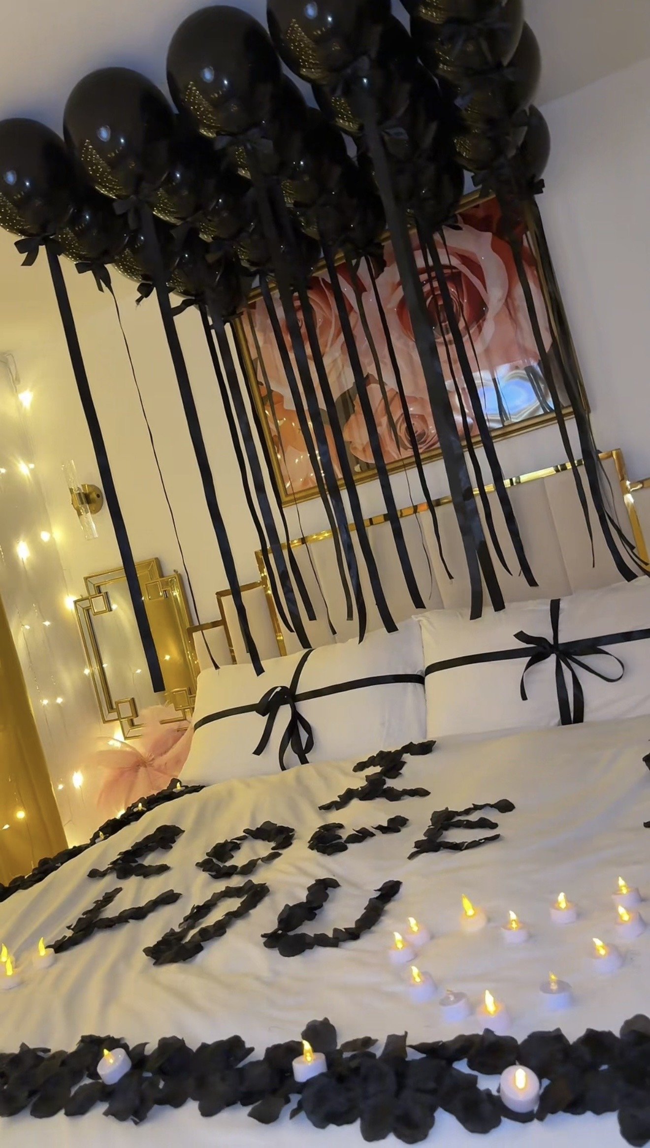 Decorated bed with pillows tied with black ribbons, rose petal letters spelling 'LOVE' on the bed, surrounded by black rose petals, lit candles, black balloons overhead tied with black ribbons, and string lights on the wall, creating a romantic setting.