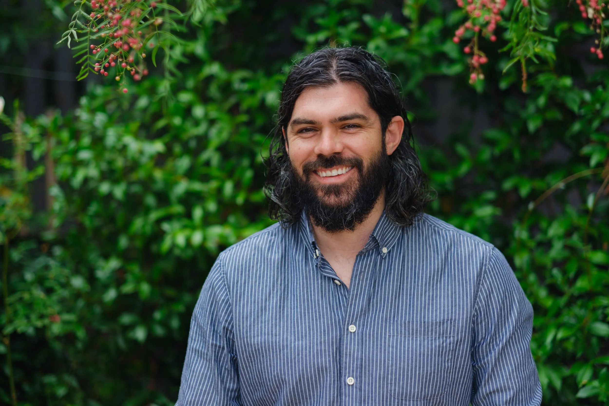 A smiling man with long dark hair and a beard, wearing a blue striped button-up shirt, standing in front of green foliage with pink berries.