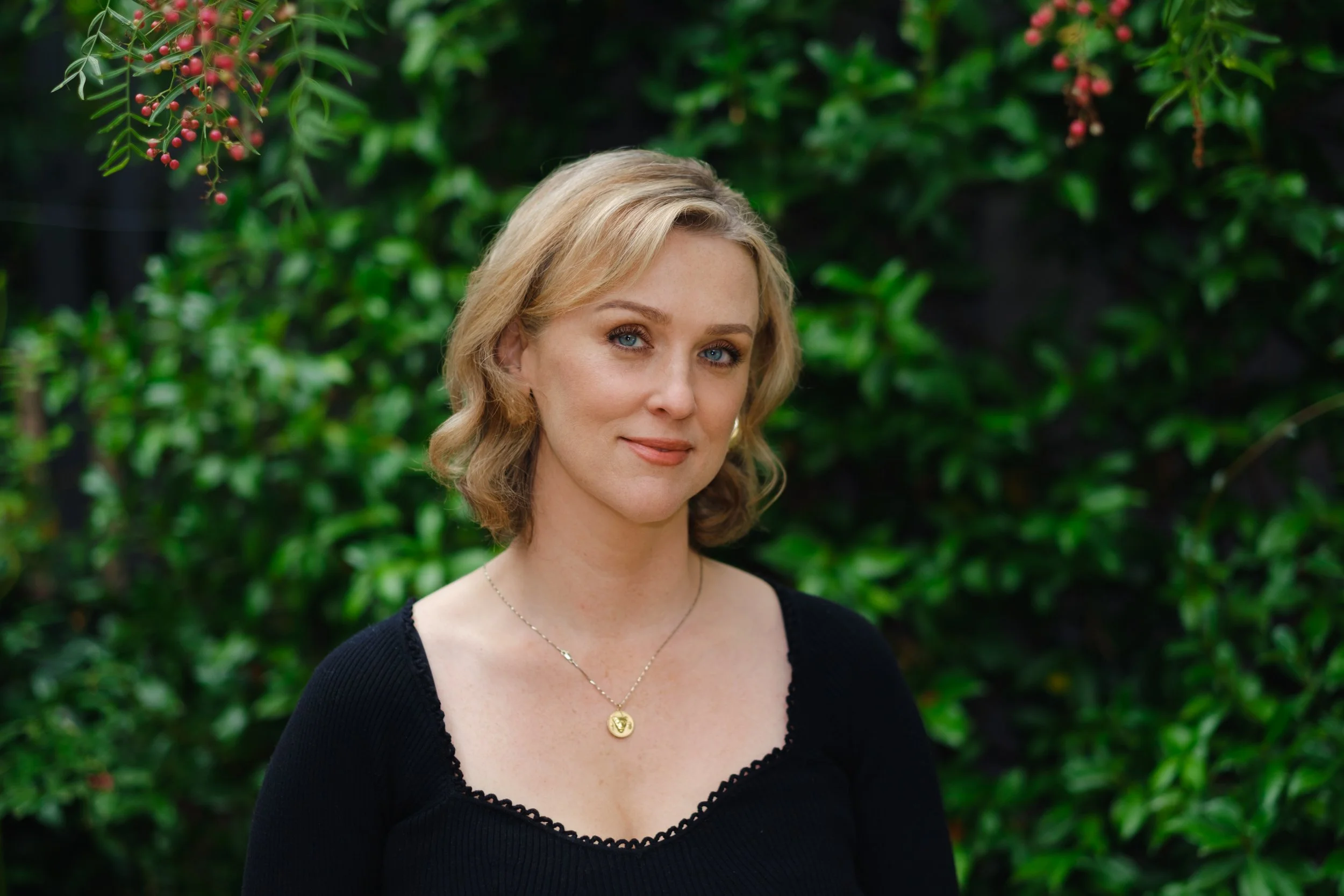 Portrait of a woman with blonde hair and blue eyes standing outdoors with greenery in the background.