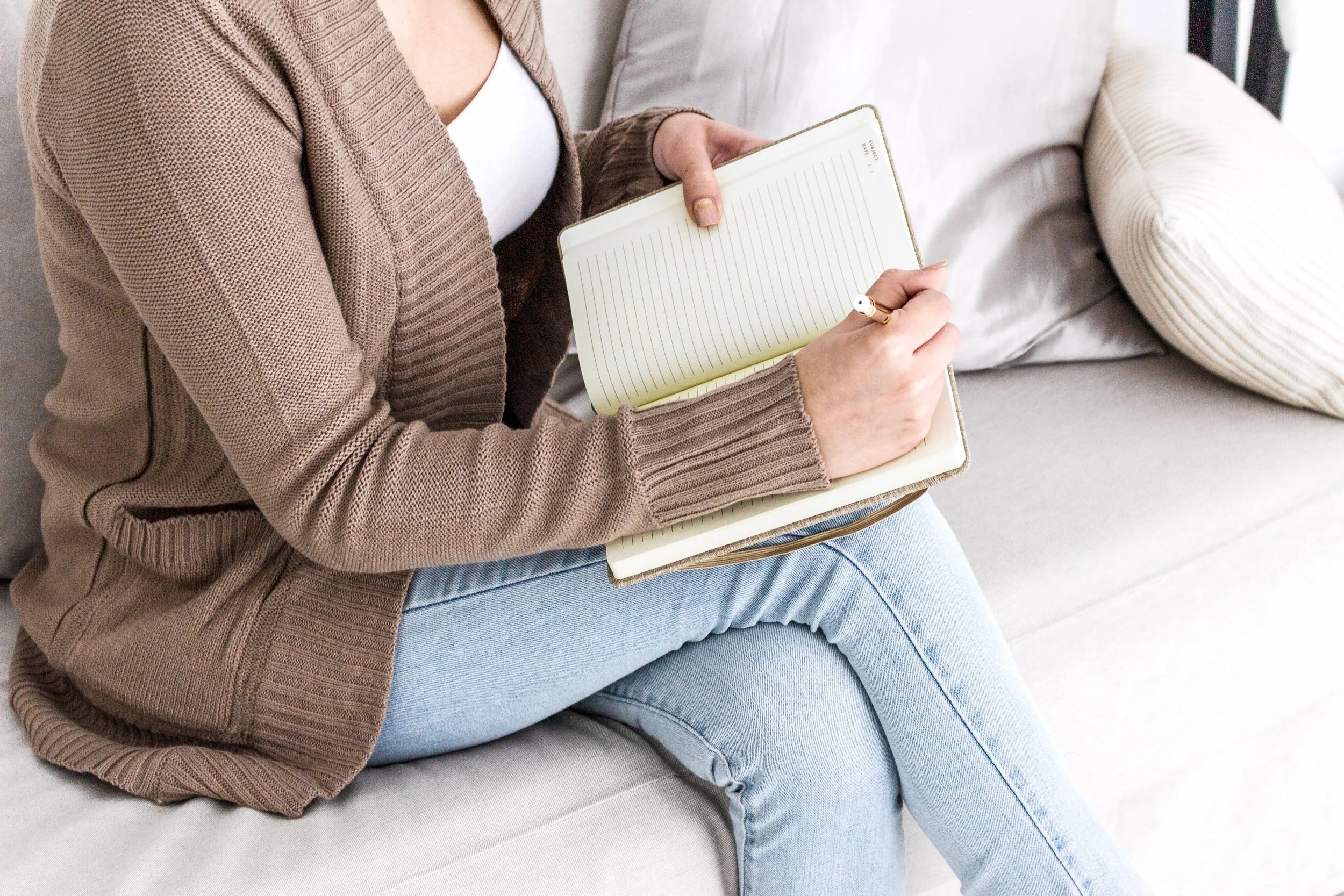 Person sitting on a sofa holding an open blank notebook with lined pages, wearing a brown cardigan and light blue jeans.
