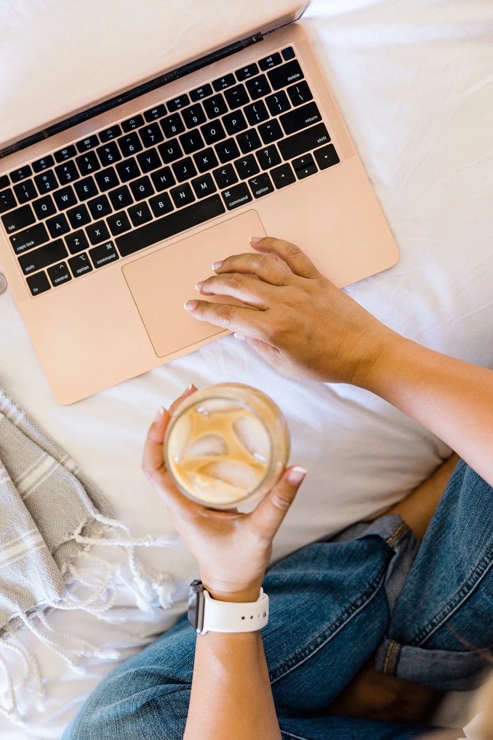 A person using a pink laptop with a hand on the keyboard, holding a glass of iced coffee, wearing a white smartwatch, sitting on a bed next to a beige and white striped blanket.