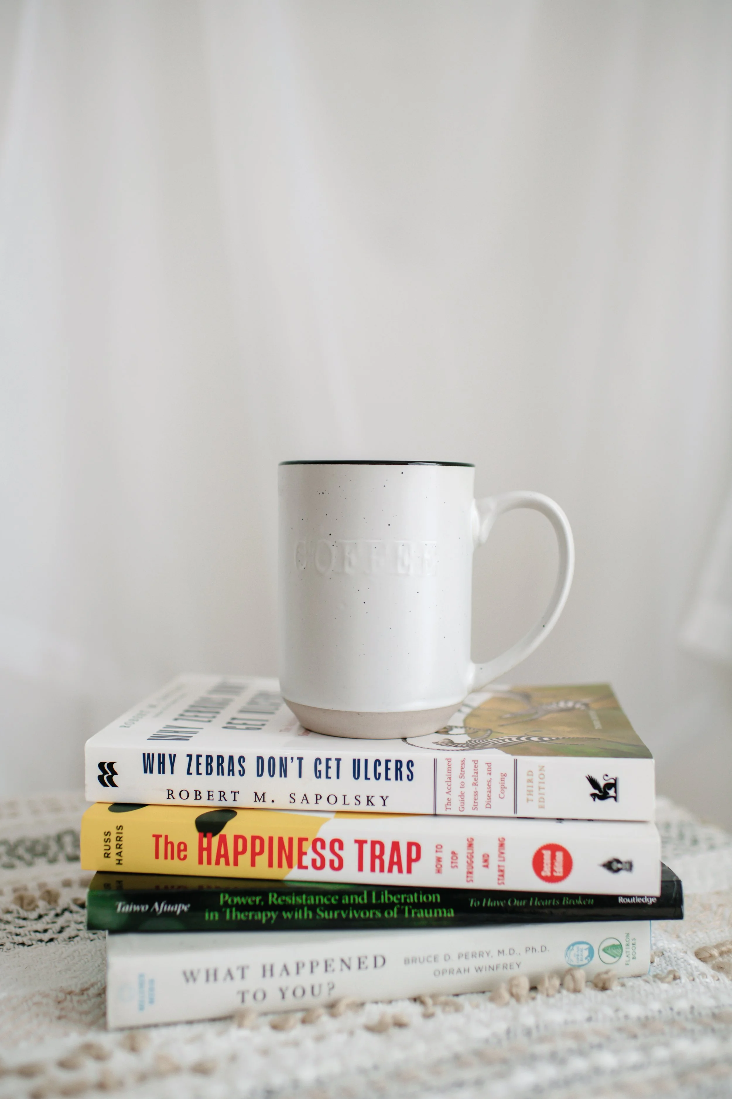 Stack of four books with a white mug on top, placed on a crocheted table covering against a white background.