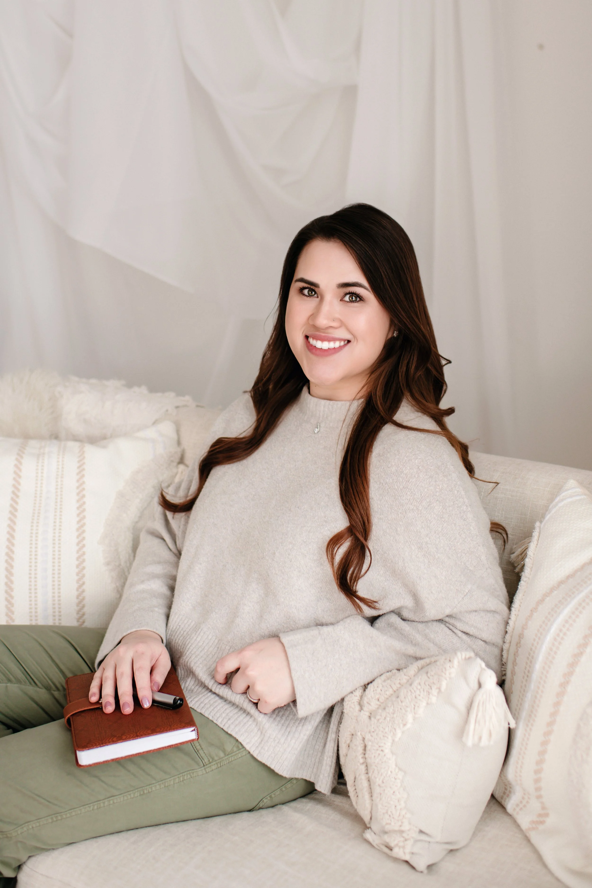 A young woman with long brown hair smiling while sitting on a beige sofa, holding a brown notebook and a black pen, in a cozy living room.