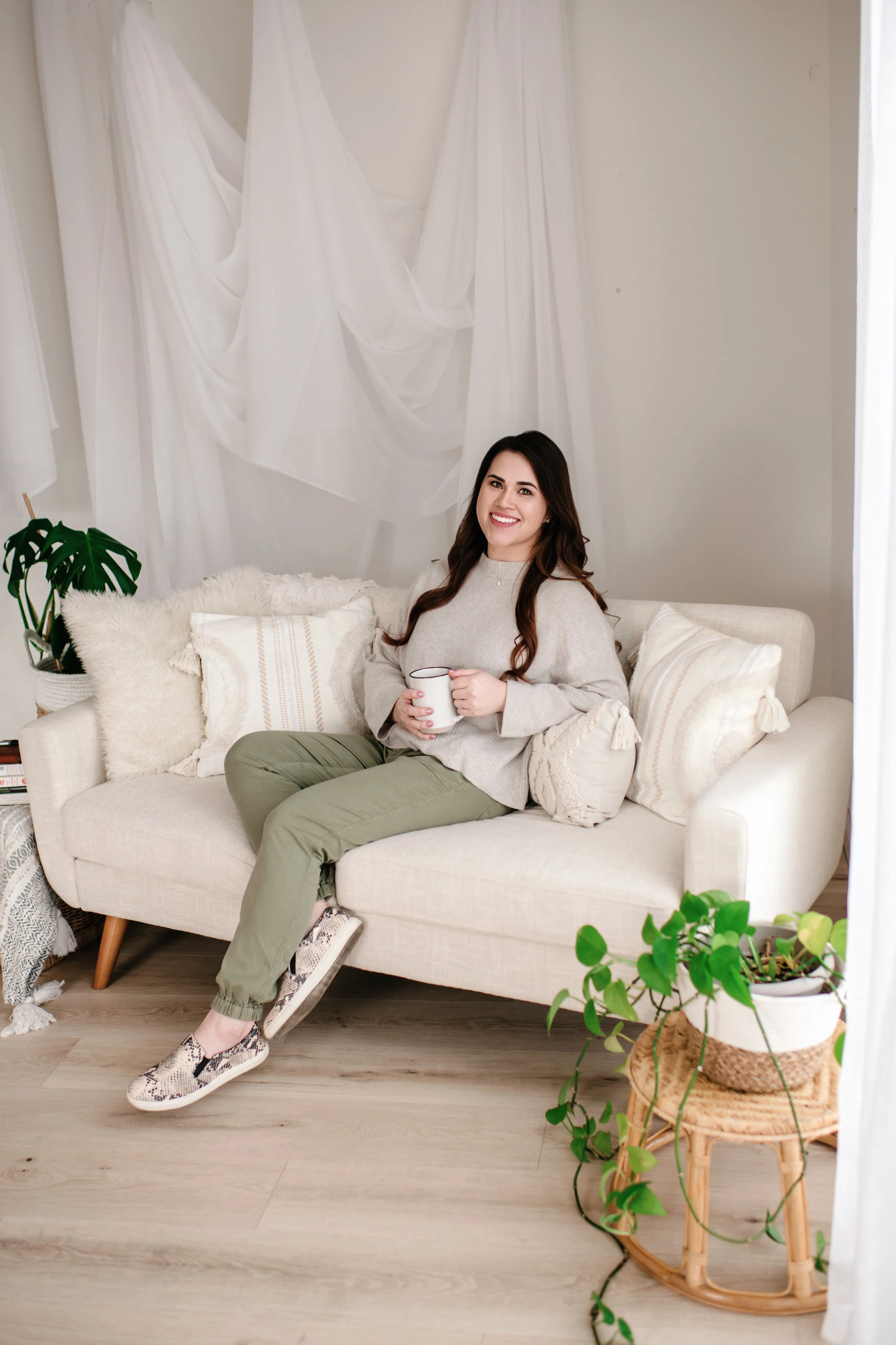 A woman with long dark hair smiling and sitting on a cream-colored sofa, holding a white mug, in a cozy living room with white drapes and indoor plants.
