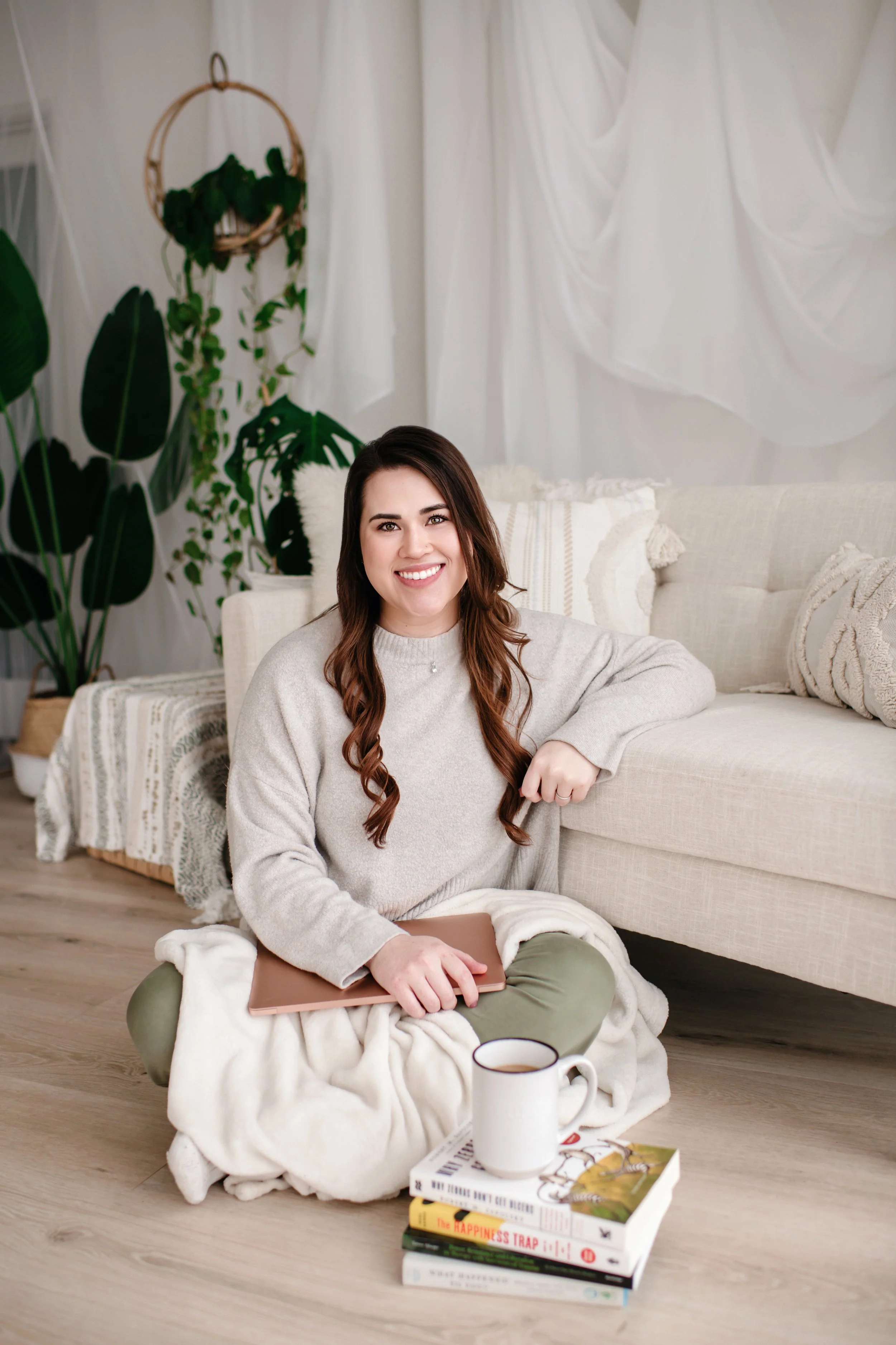 A woman with long brown hair, wearing a beige sweater, sitting cross-legged on the floor with a blanket on her lap, smiling, with a laptop in her hand. She is in a cozy living room with green plants and white curtains.