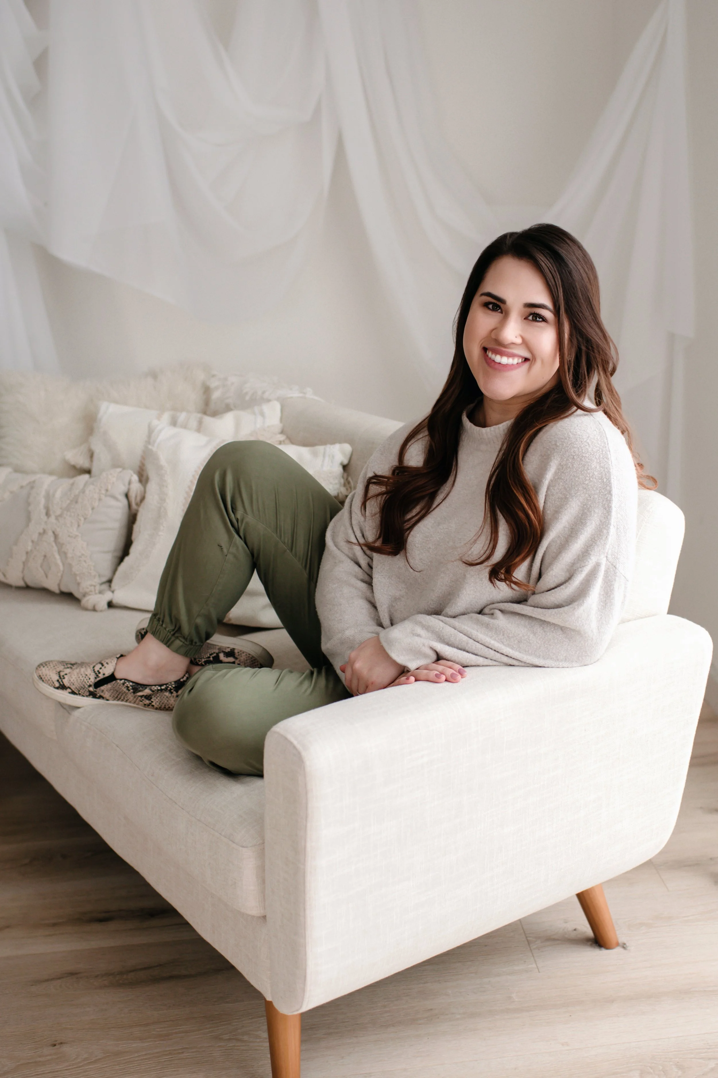 A young woman with long brown hair, wearing a beige sweater and green pants, sitting on a white sofa with a neutral expression, in a cozy room with cream-colored walls and decorative pillows.