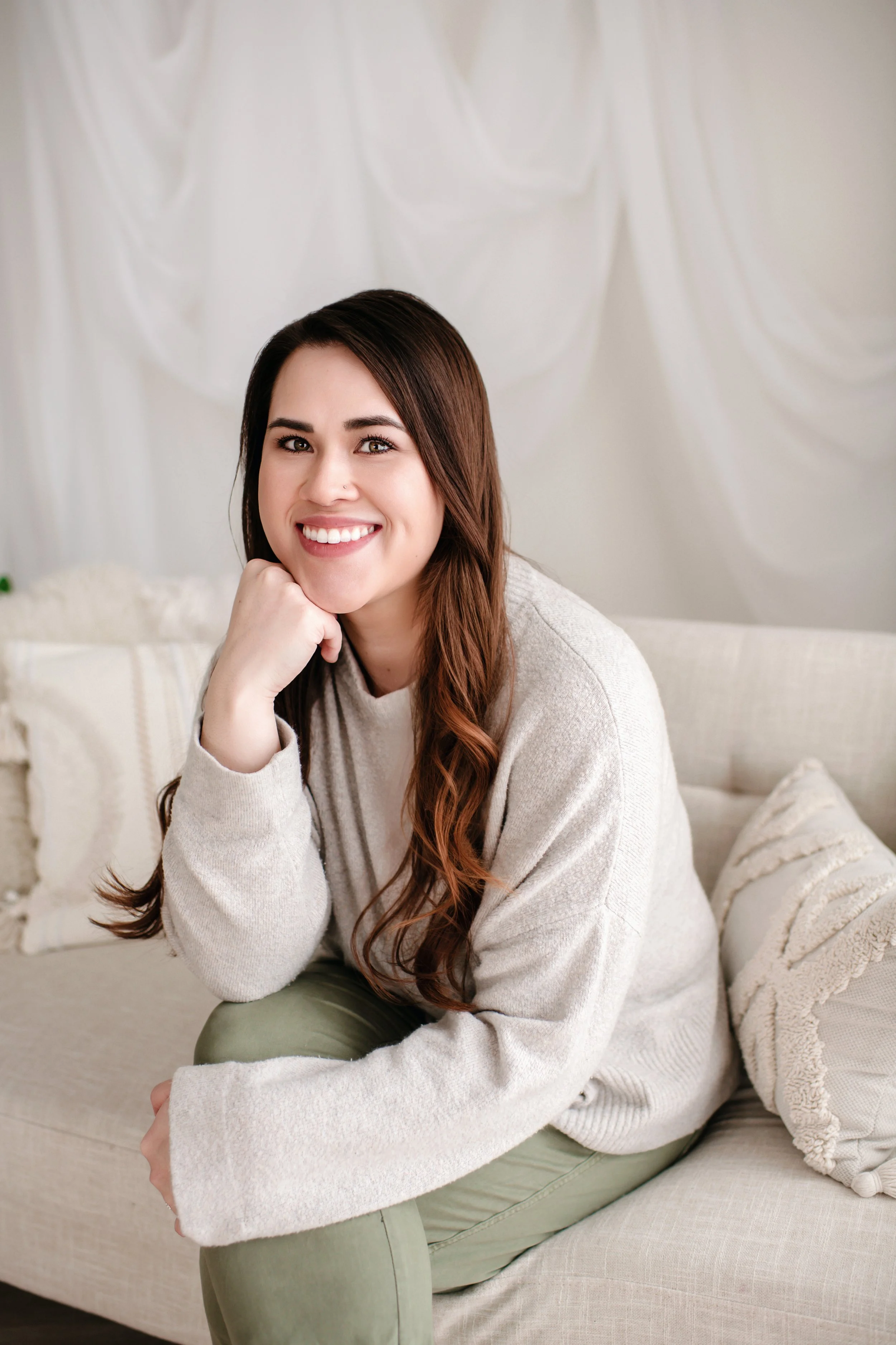 A young woman with long brown hair, smiling, sitting on a light-colored sofa in a cozy, well-lit room.