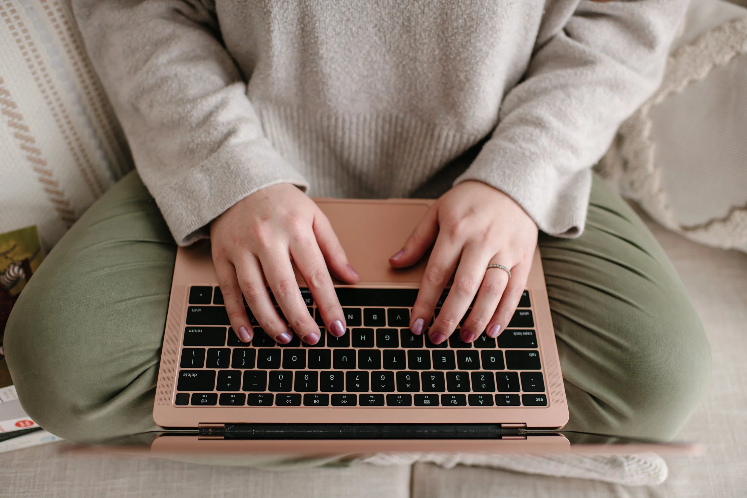 Person sitting cross-legged on a beige and cream-colored couch, using a rose gold laptop with black keyboard.