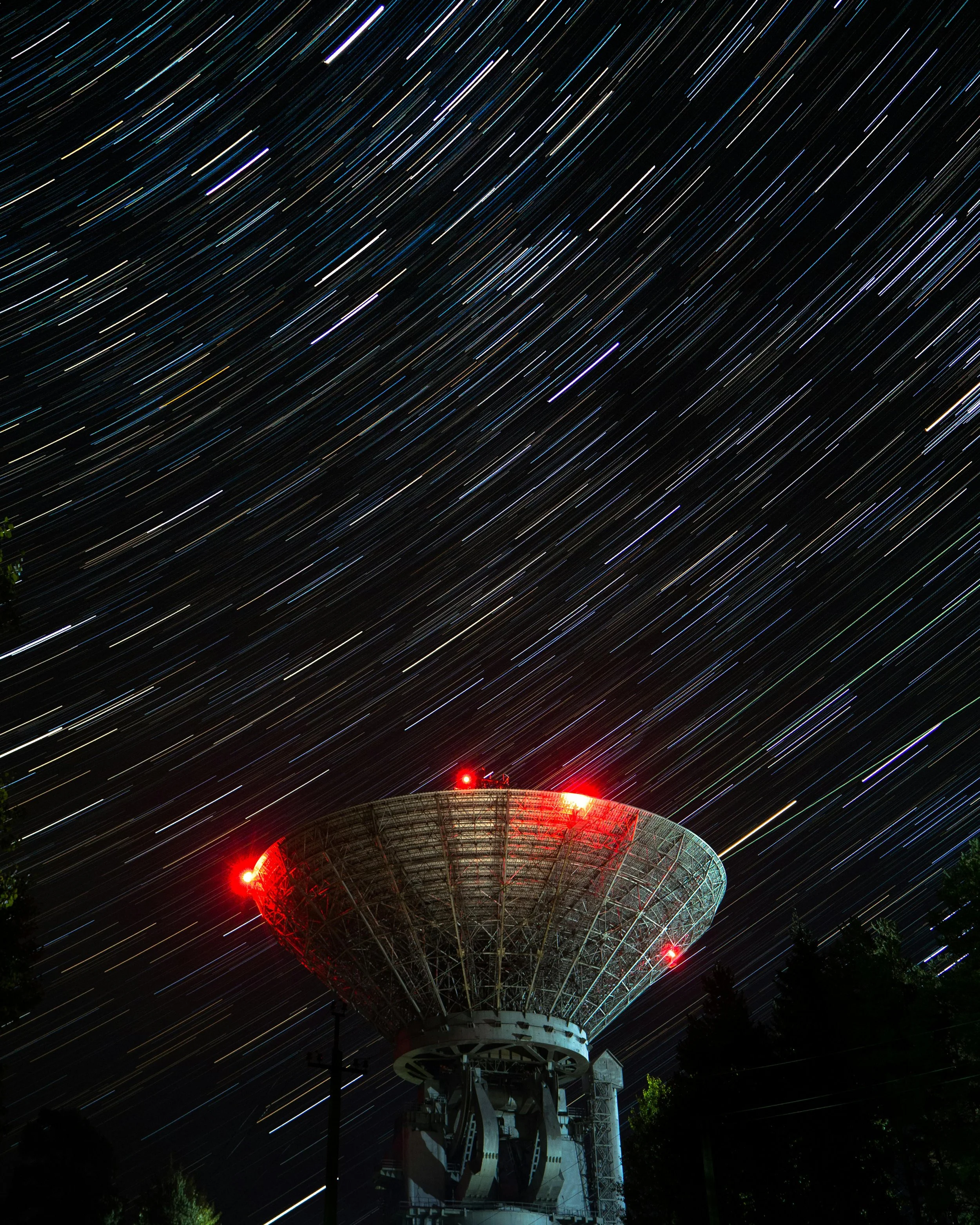 Time-lapse photo of star trails in the night sky over a large radar or communication structure with red lights
