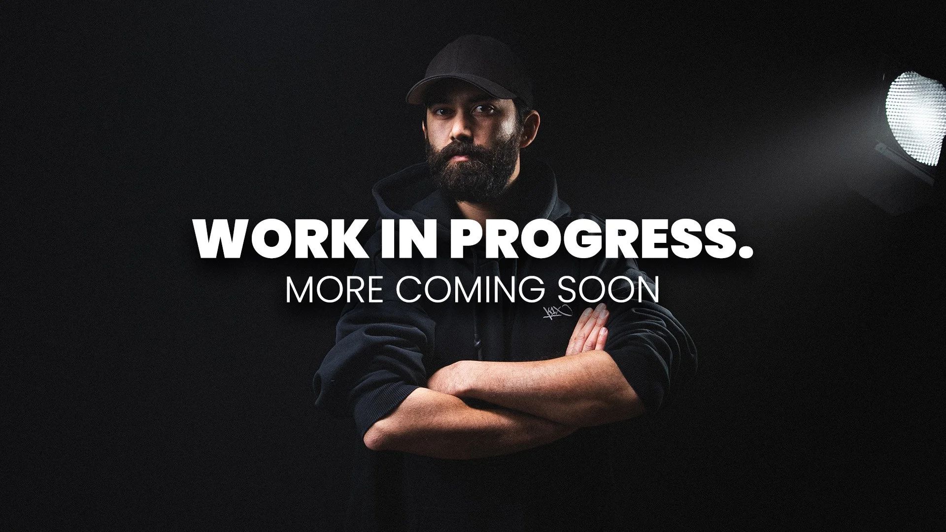 Ein Mann mit dunklem Outfit und Kappe steht vor einem schwarzen Hintergrund, mit Text 'WORK IN PROGRESS. MORE COMING SOON' darüber.