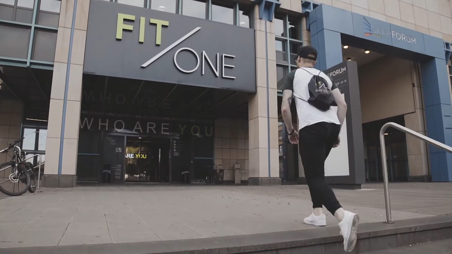 Mann betritt Fitnessstudio vor einem modernen Gebäude mit Fahrrad und Fahrradbügeln draußen.