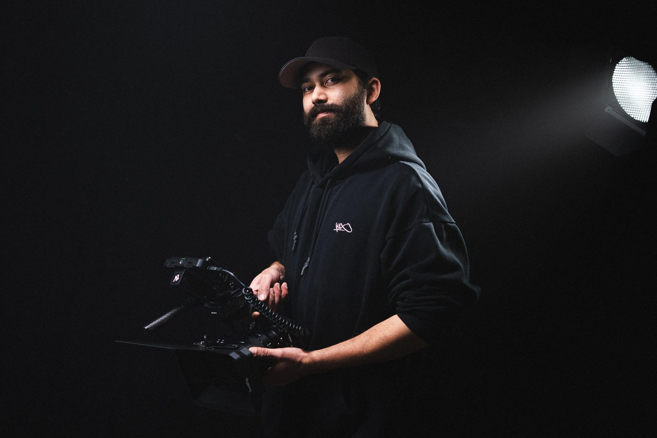 Ein Mann in schwarzer Kapuzenjacke und schwarzem Cap hält eine Videokamera auf dunklem Hintergrund.