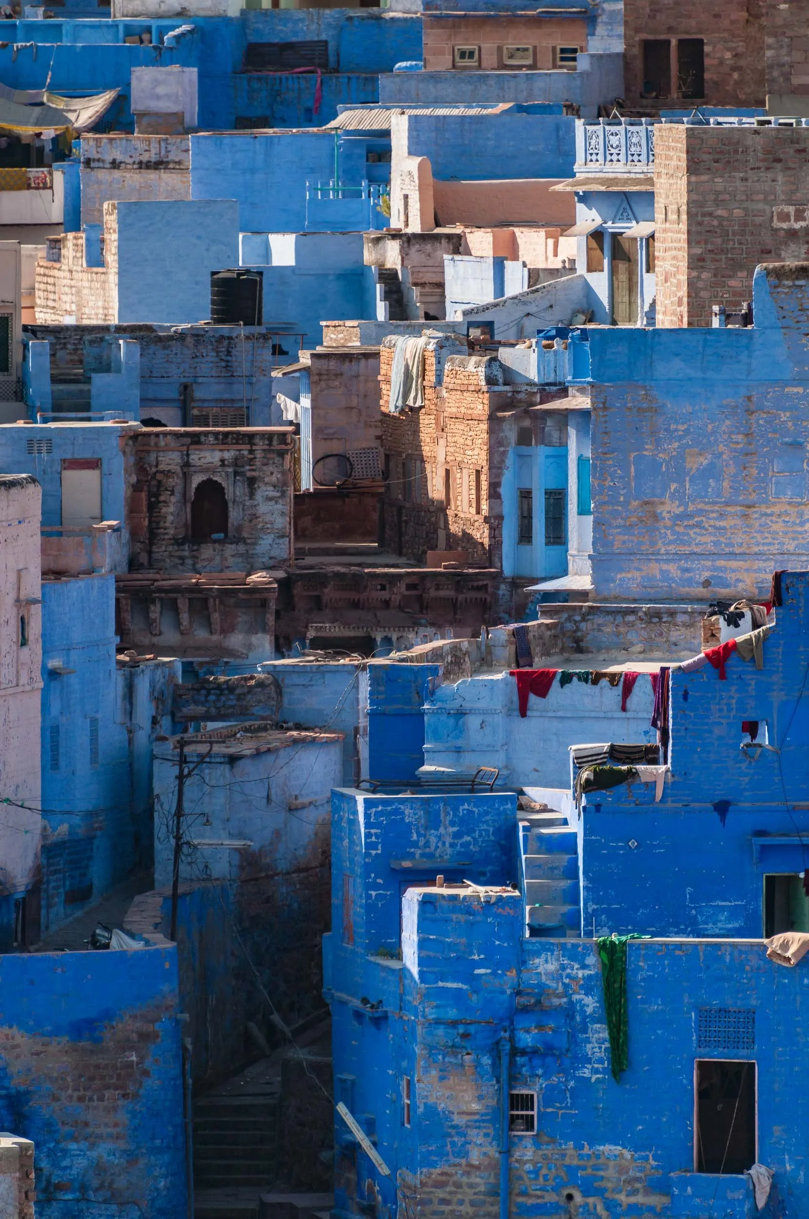 A landscape of densely packed houses painted in various shades of blue, with some exposed brick walls, laundry hanging on clotheslines, and stairs connecting different levels.