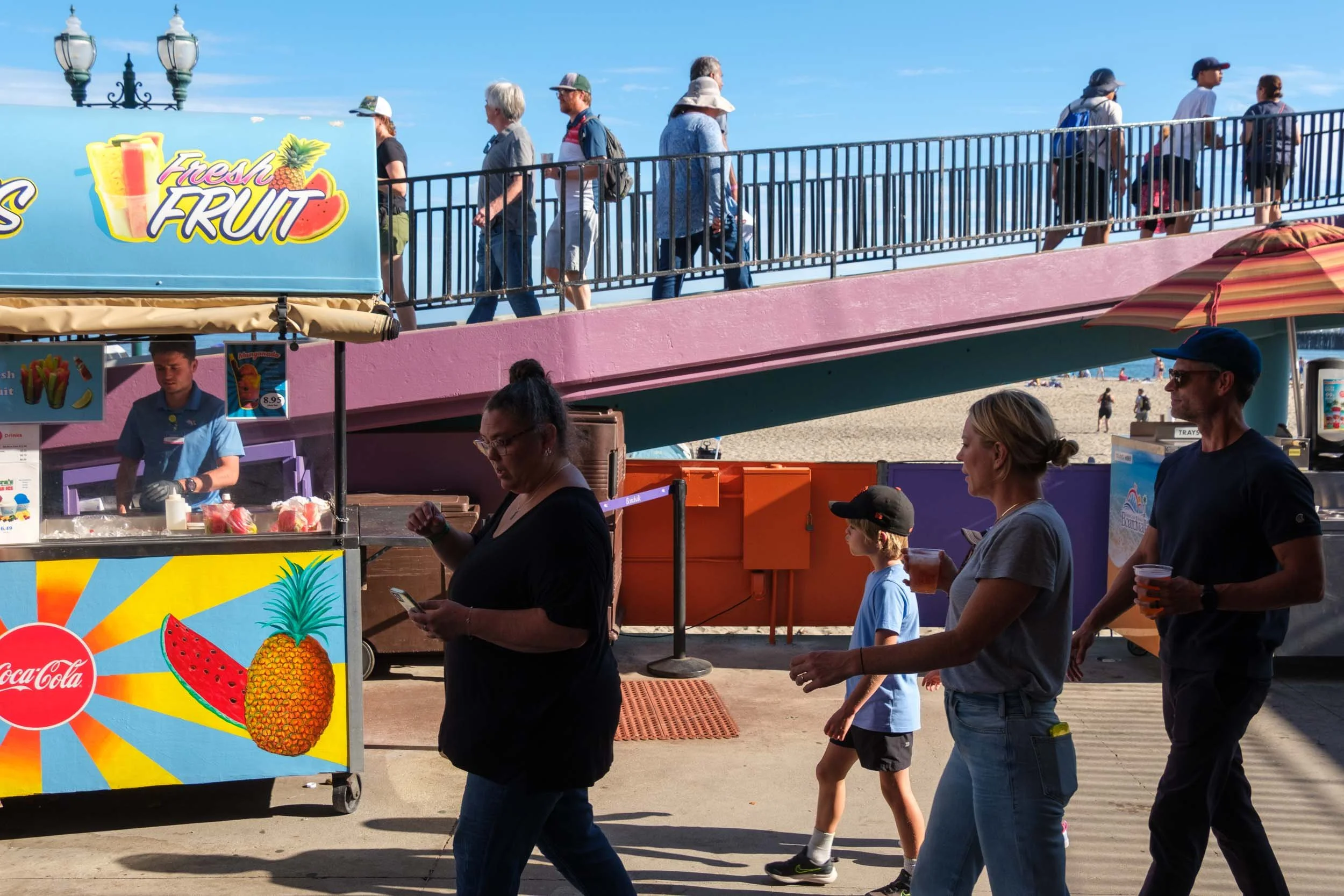 People walking along a boardwalk at the beach with a food stand selling fresh fruit. A sign features a pineapple and a watermelon. Some people carry drinks and wear casual summer clothing.