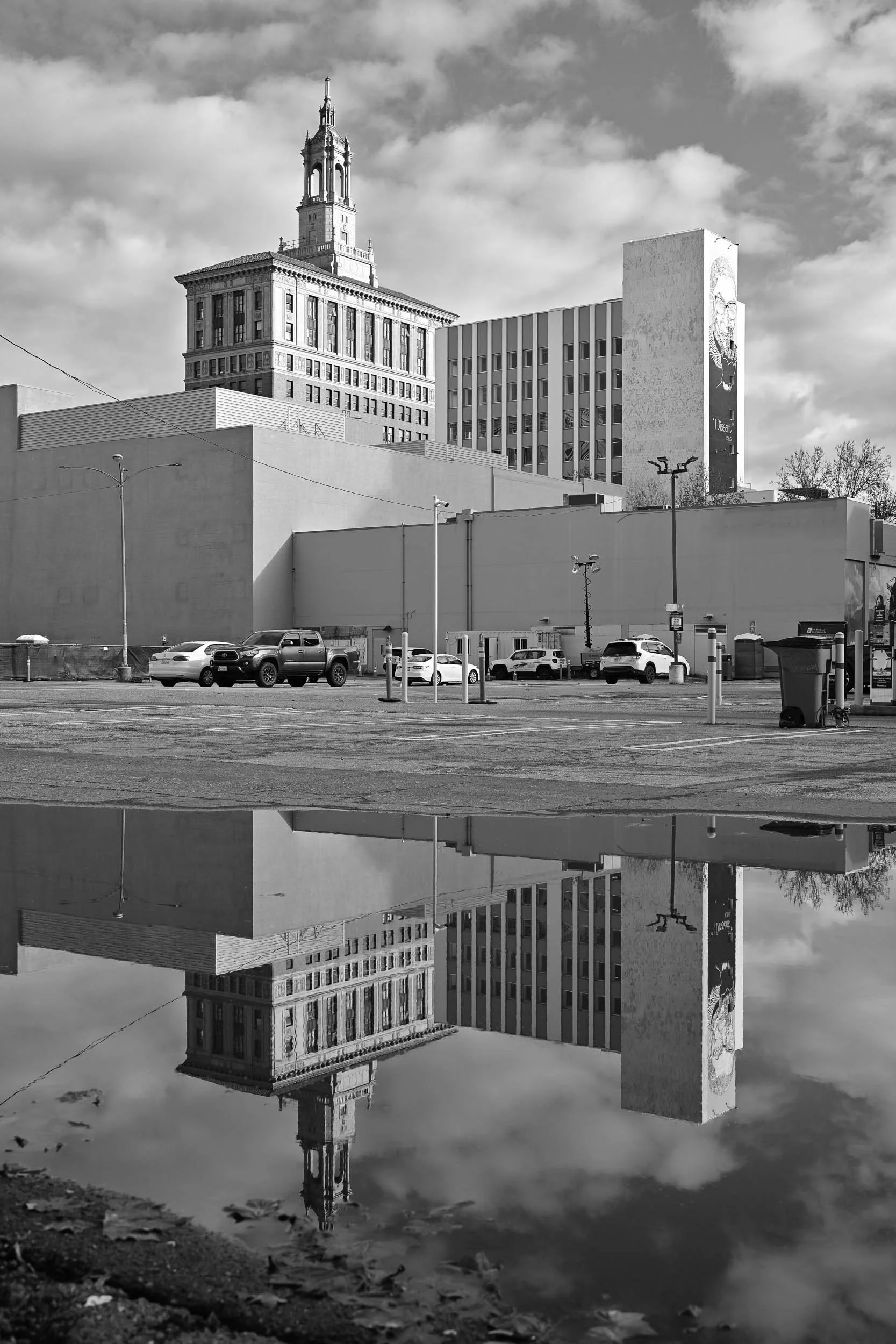 A cityscape with tall buildings, cars parked in a lot, and a reflection of the buildings in a puddle on the ground.