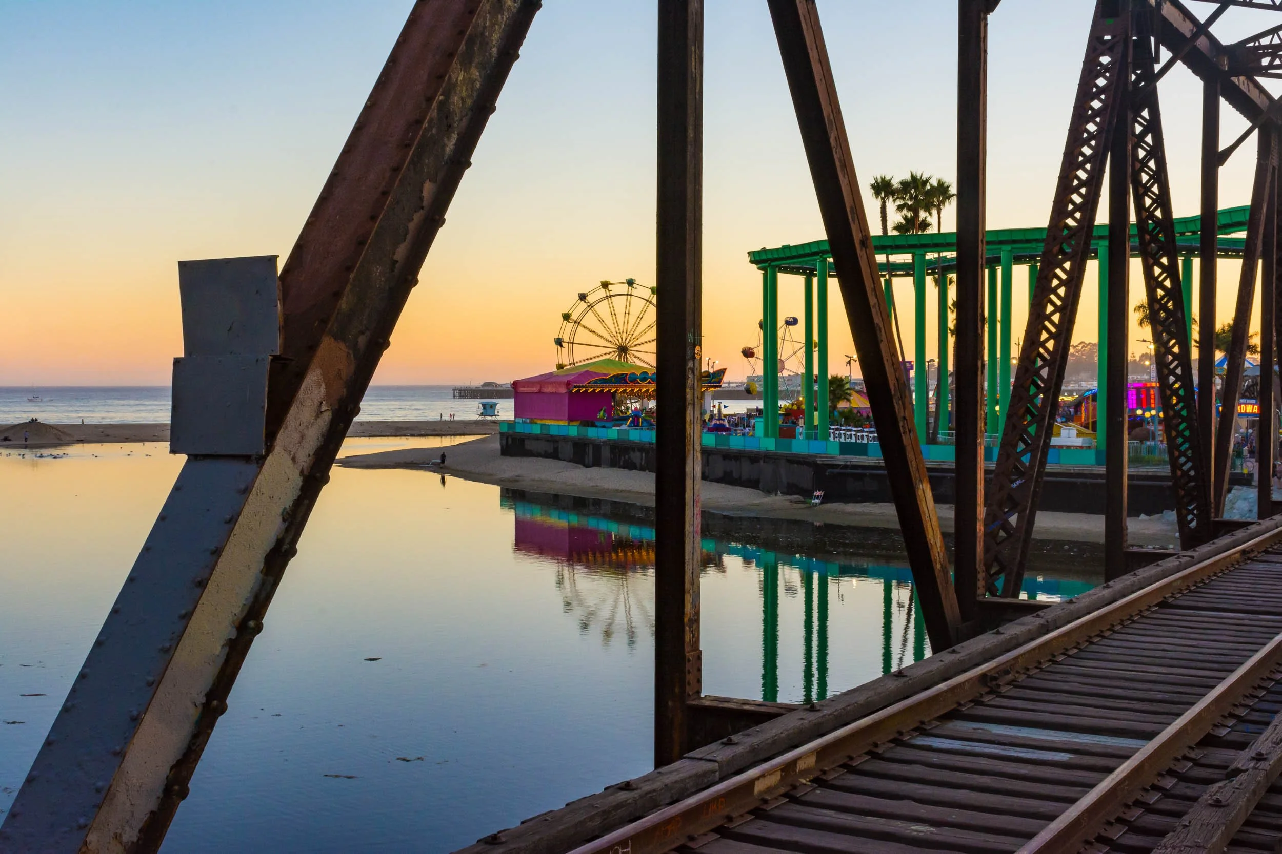 View of a deserted amusement park on a beach at sunset, seen through the rusted steel support beams of a roller coaster track, with a Ferris wheel, colorful rides, and palm trees in the background.