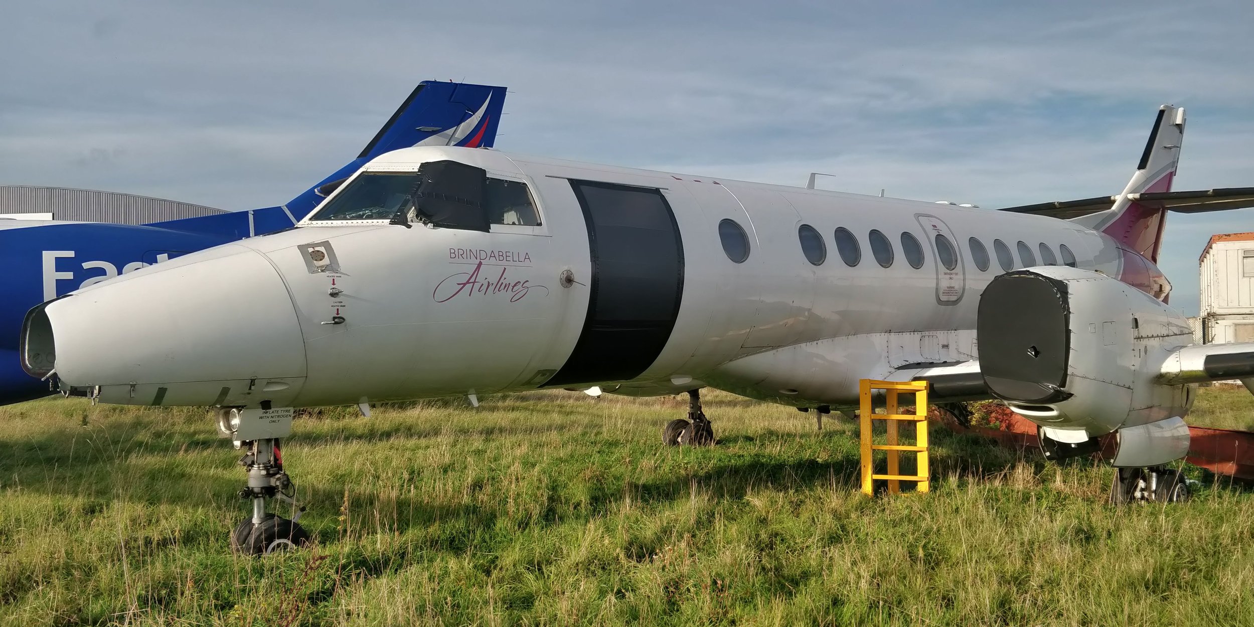 A small white airplane with 'Brindabella Airlines' written on the side, parked on grass with a yellow step ladder nearby, next to a larger FedEx cargo plane in the background.