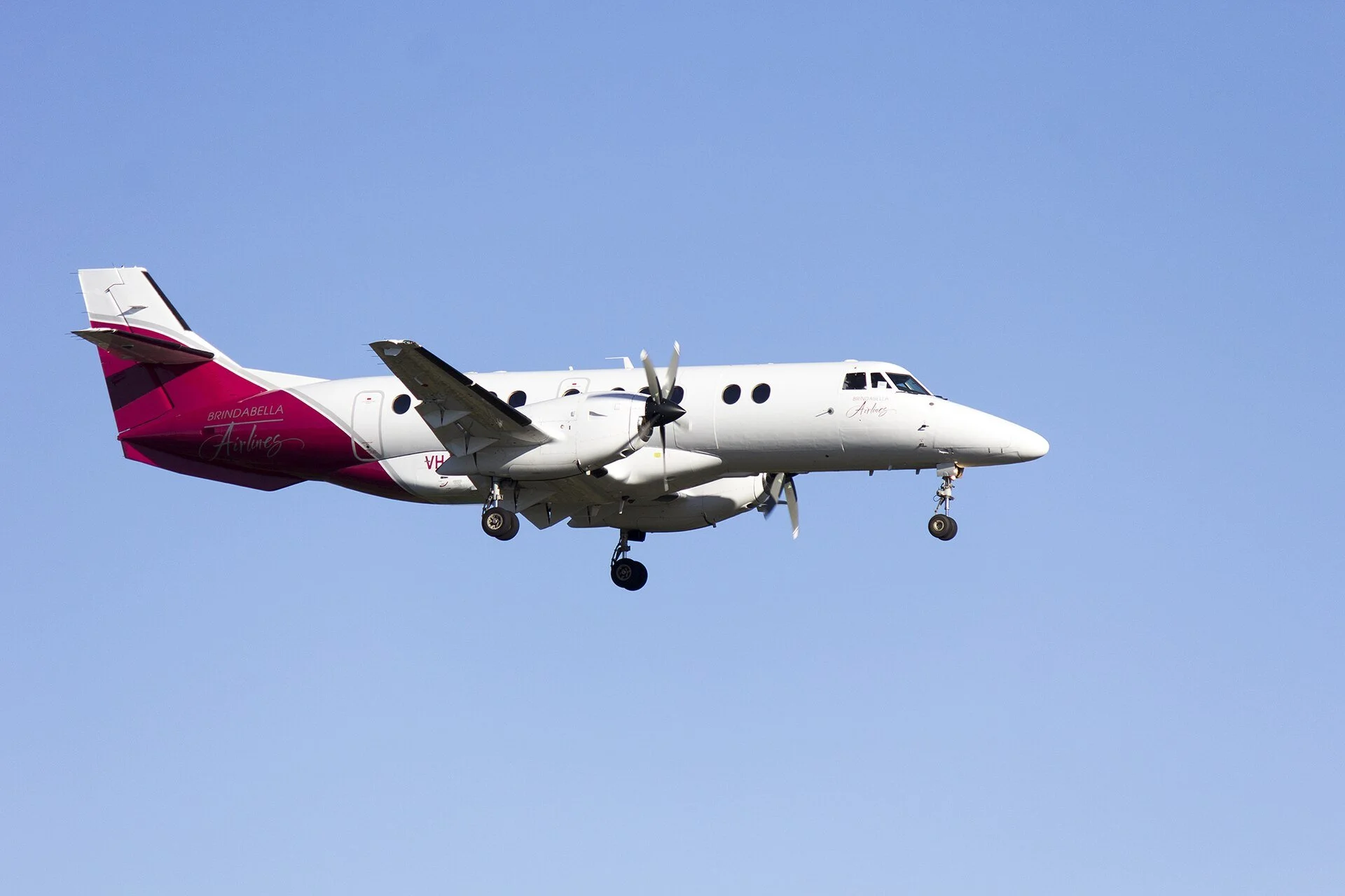 A white and pink twin-engine propeller airplane flying in a clear blue sky with extended landing gear.
