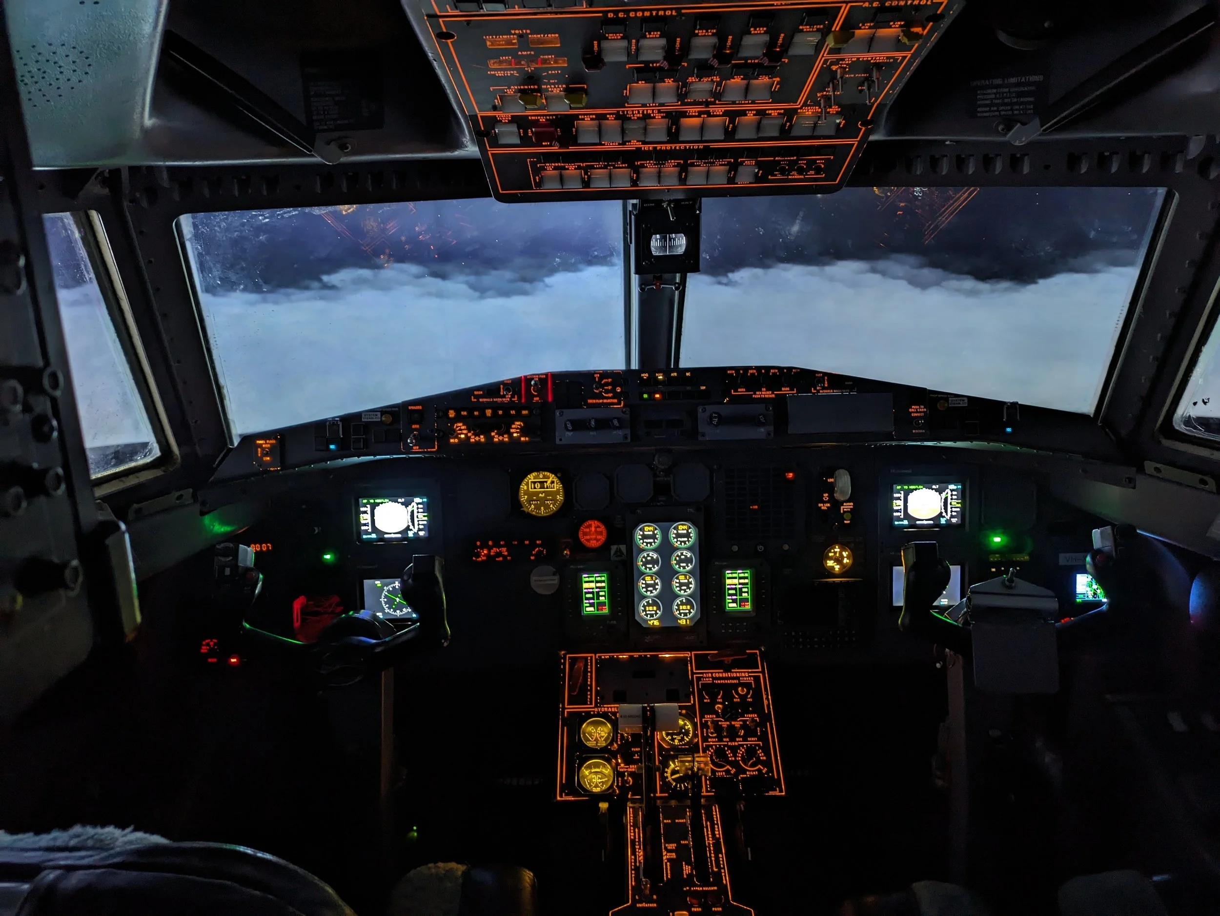 Cockpit of an airplane with illuminated controls and gauges, showing a view of clouds and sky outside the windshield.