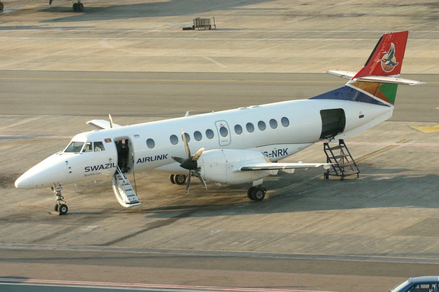 Small white airplane on airport tarmac with open door and stairs, branding indicates Swaziland Airlink, with a colorful tail logo, propellers, and passenger windows.