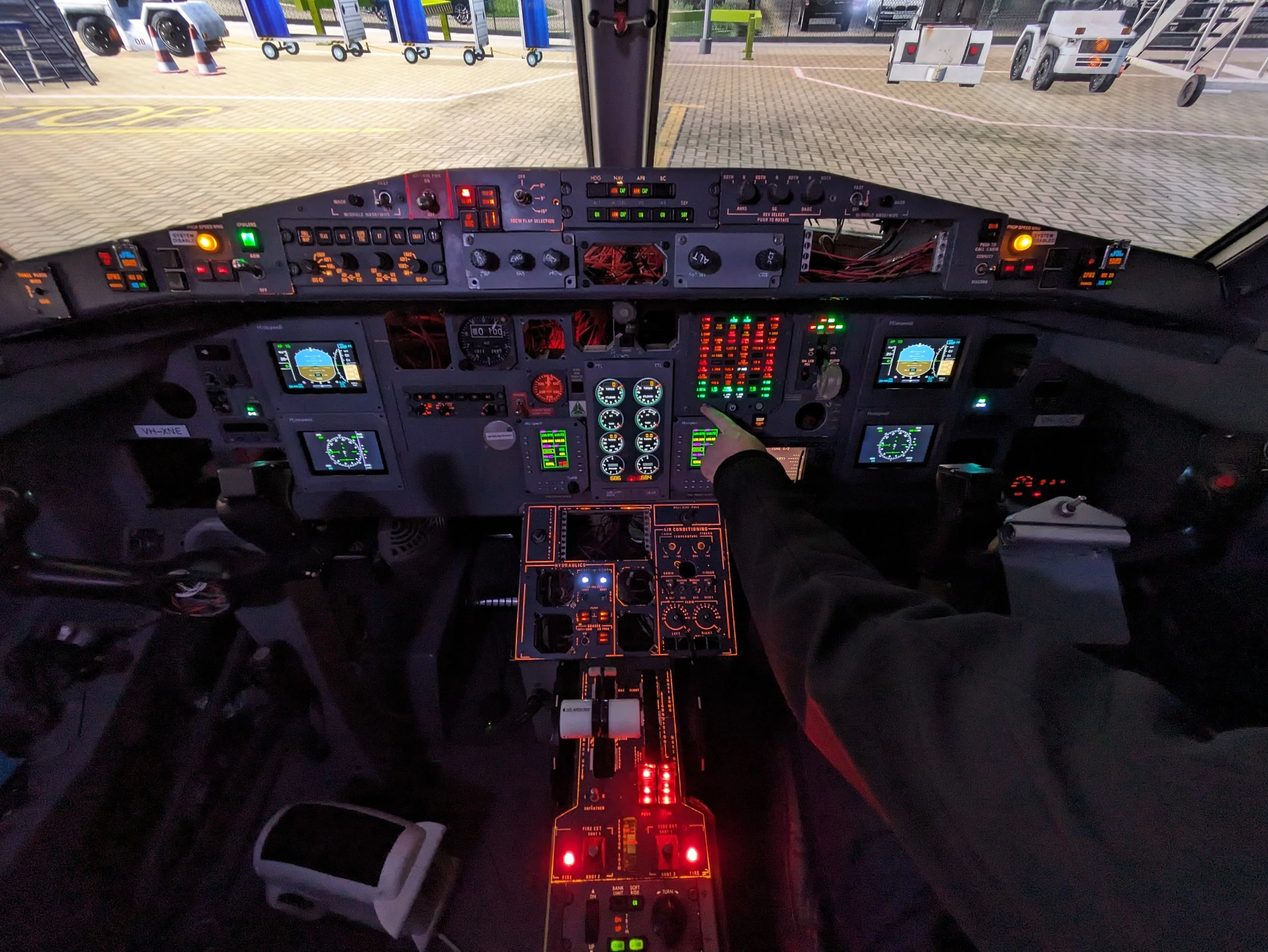 Interior view of a commercial airplane cockpit with various controls, instruments, and displays illuminated, with an airport tarmac visible through the windshield.