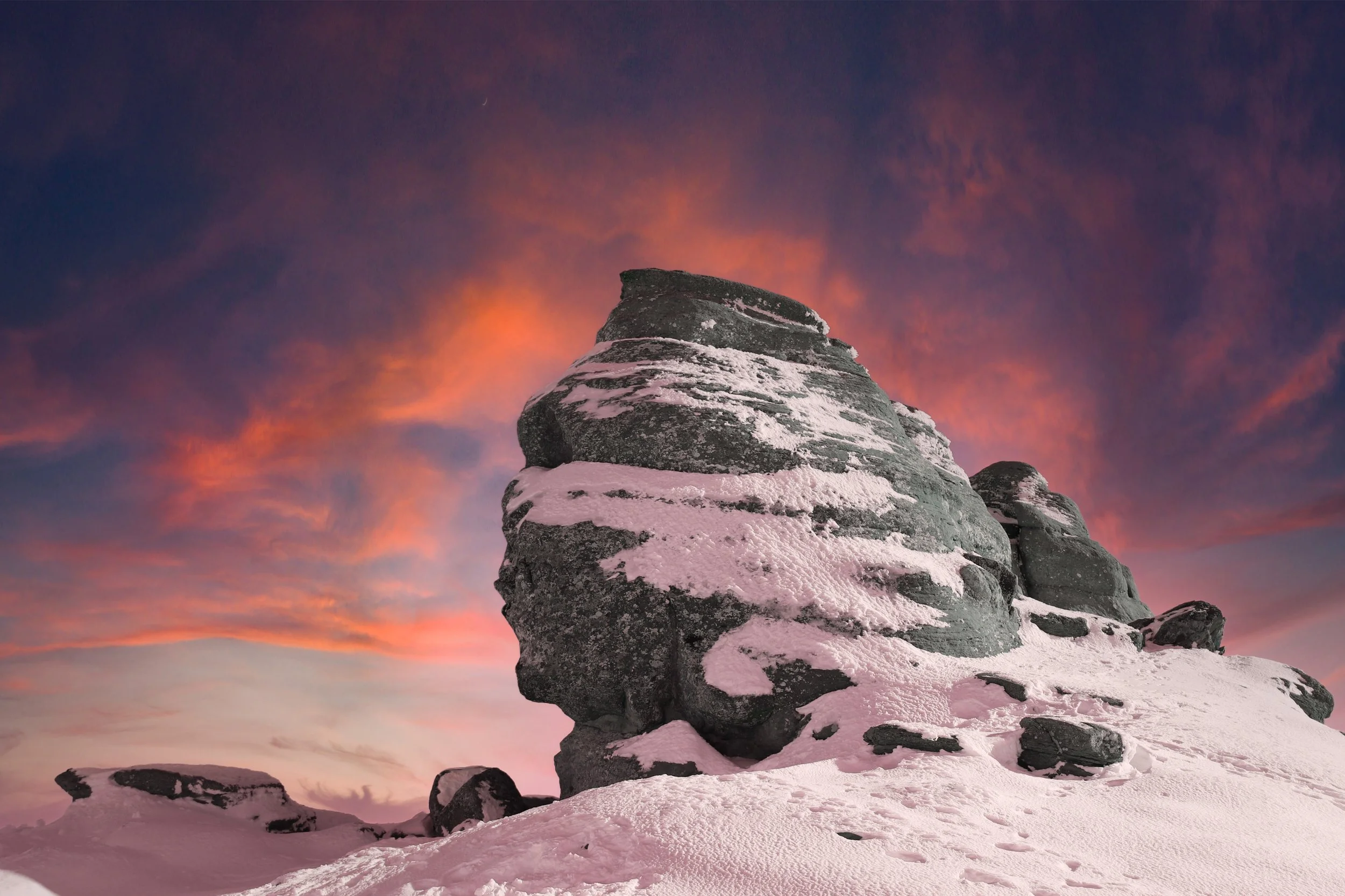 The Sphinx rock formation in the Bucegi Mountains, Romania, covered in snow under a dramatic sunset sky.