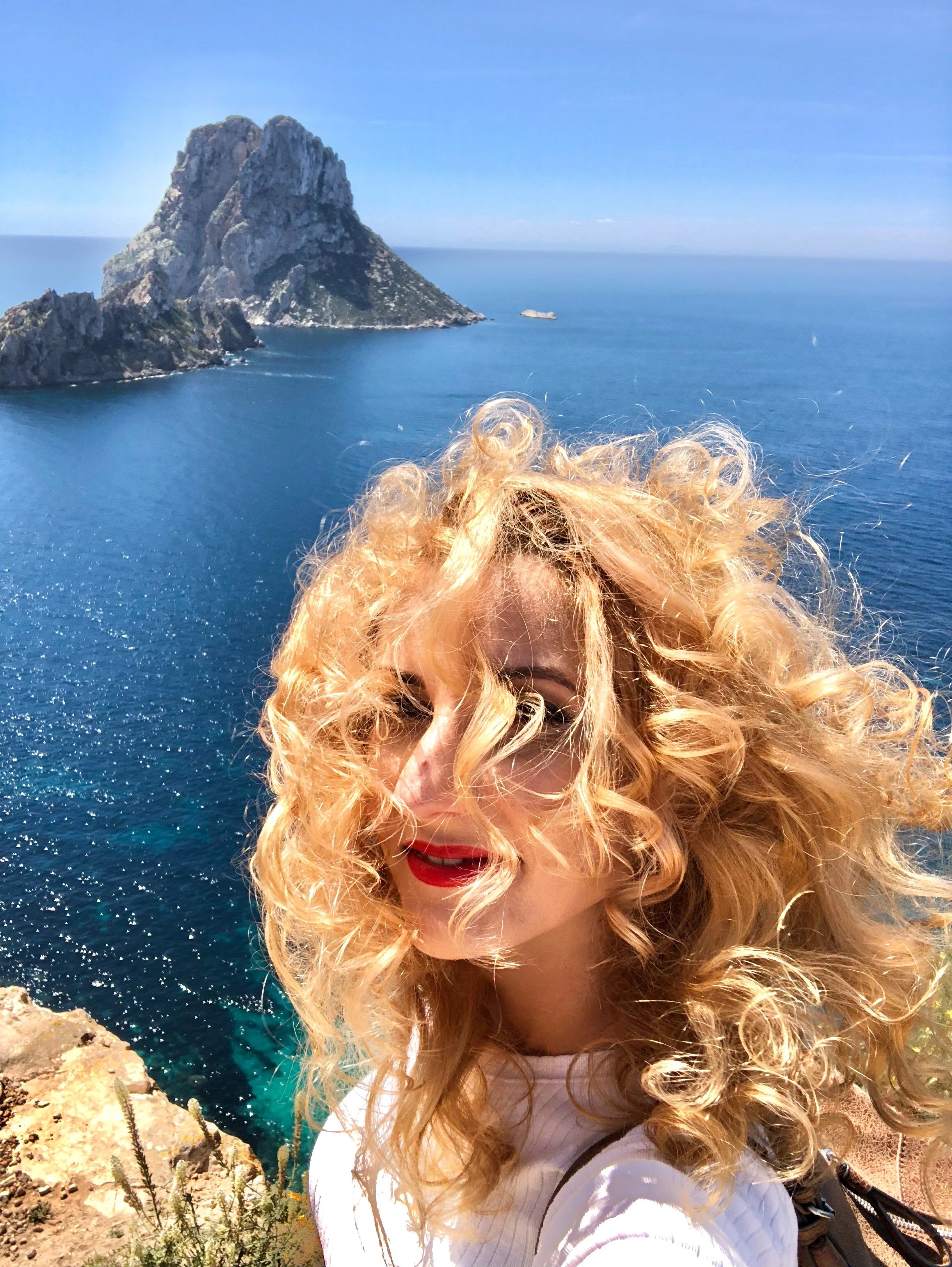 Woman with curly blonde hair taking a selfie on a cliff overlooking a turquoise cove, atlantis - ibiza