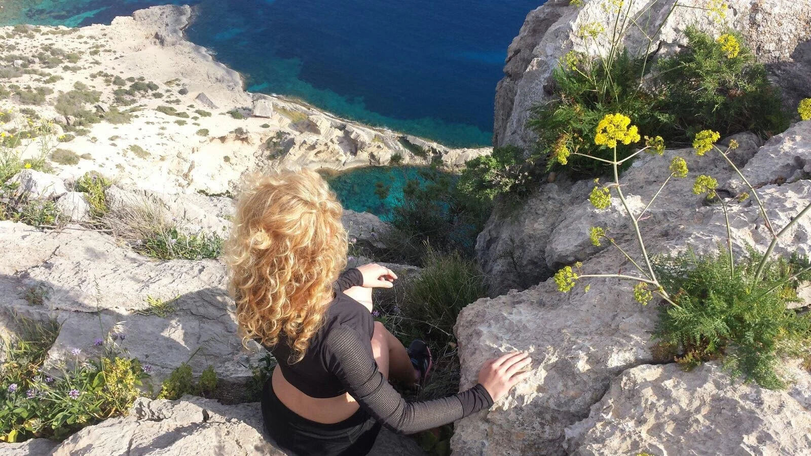 Woman sitting on rocky cliffs above a turquoise Mediterranean bay with wildflowers and limestone rocks around her in Atlantis, Ibiza