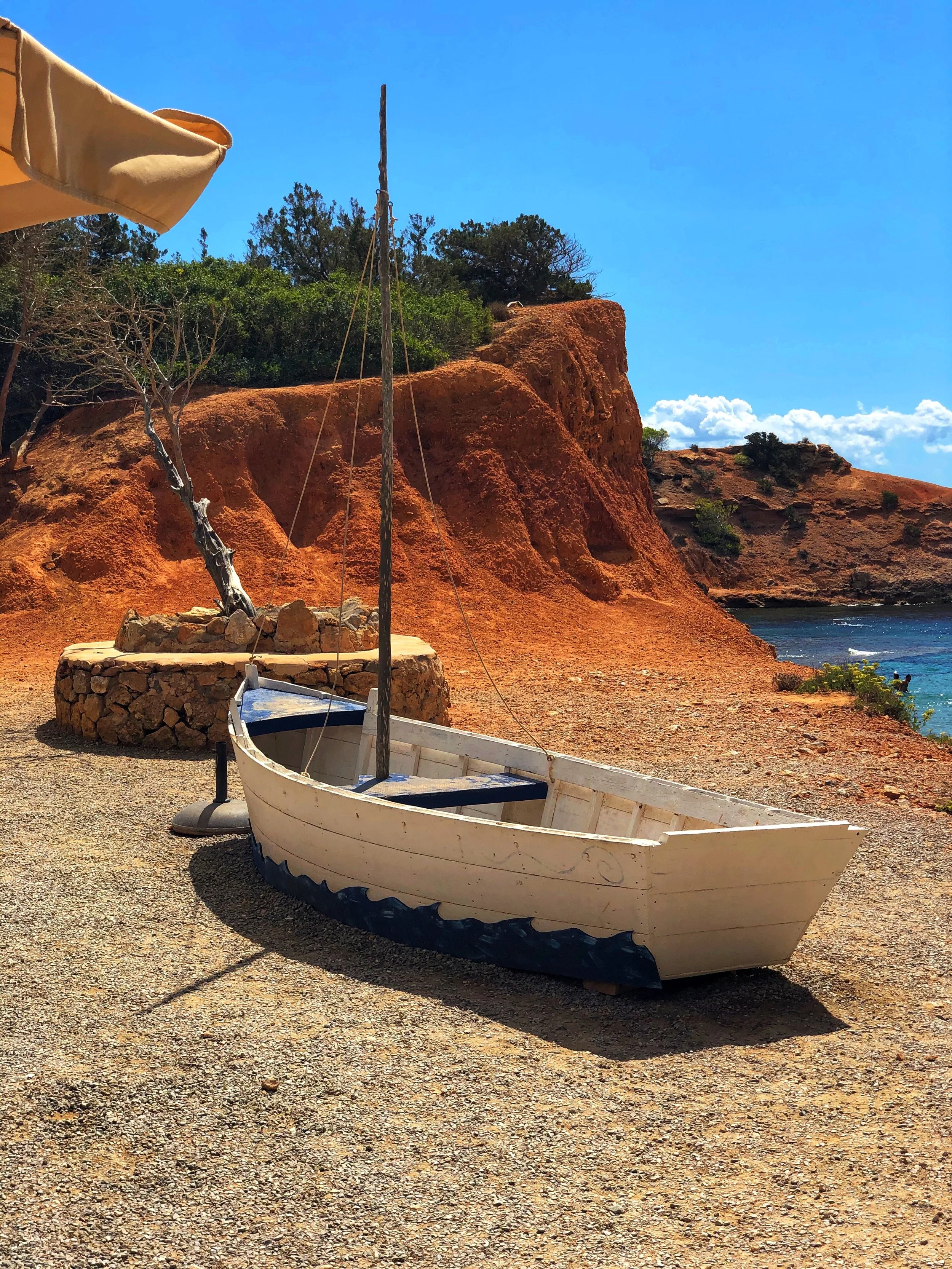 Small wooden fishing boat on a rocky beach with red cliffs and turquoise sea in Ibiza, Spain.