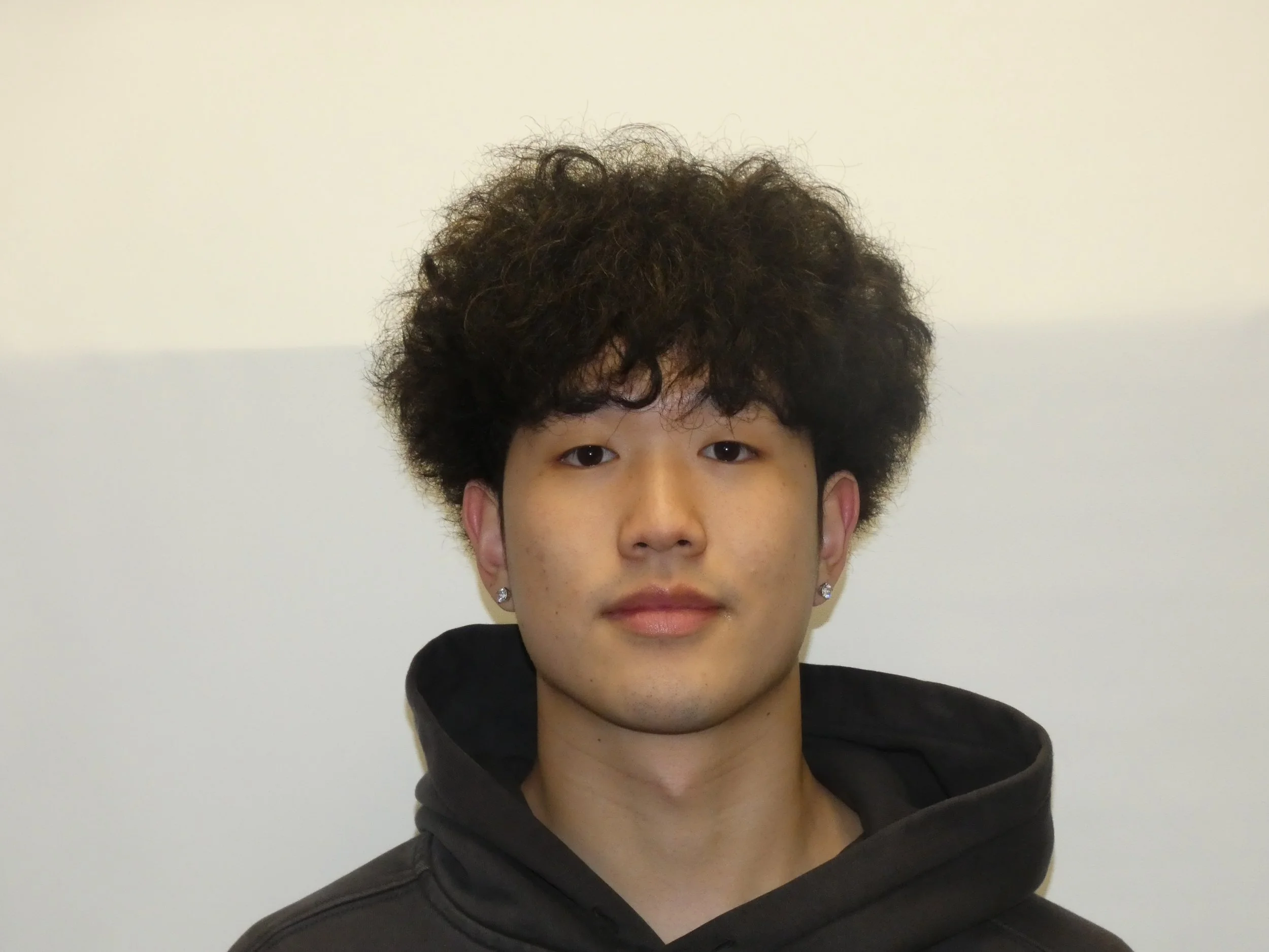 Young man with dark, curly hair, wearing earrings and a black hoodie, standing against a plain, light-colored wall.