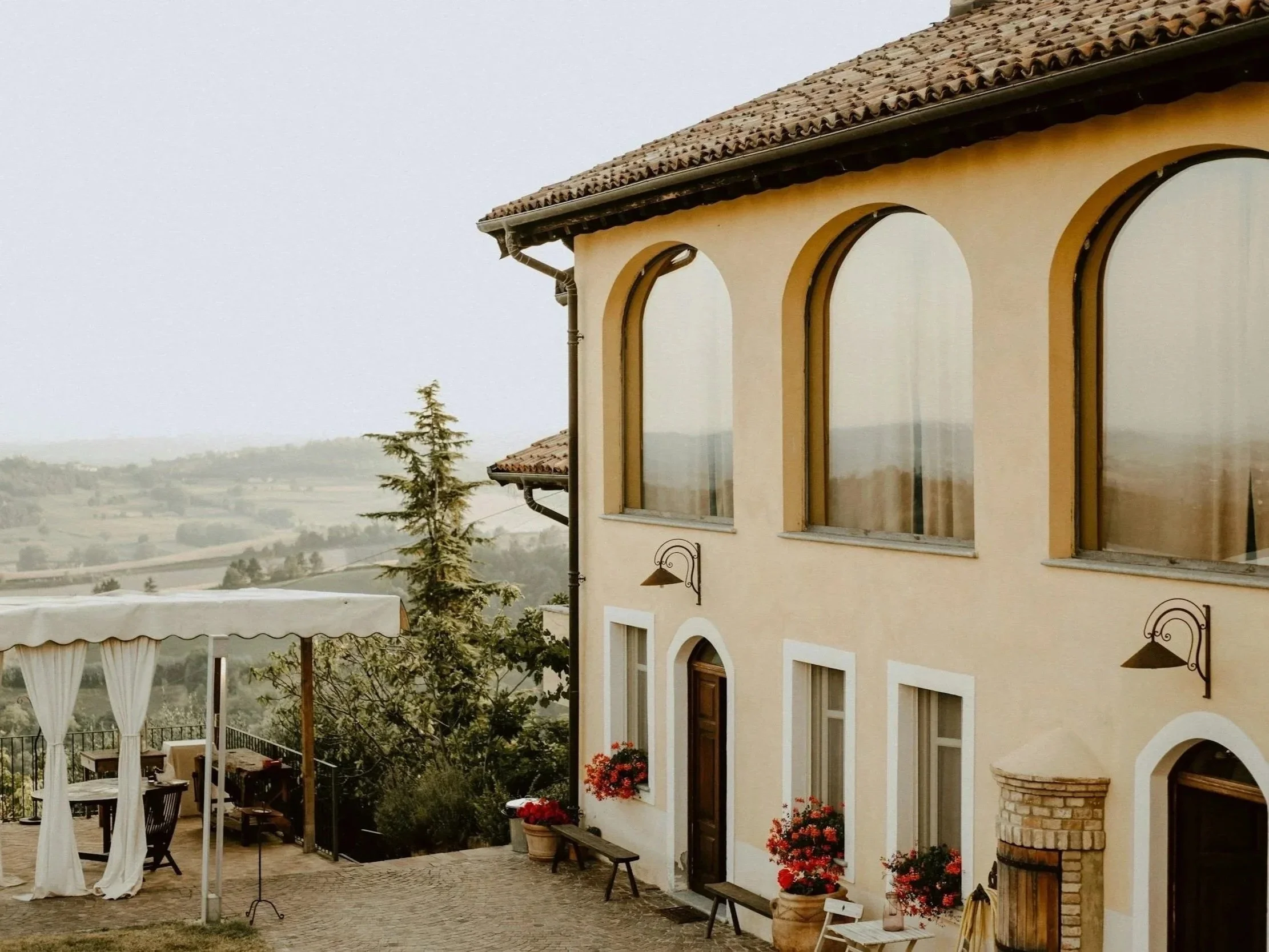 Exterior of a beige house with arched windows, red flowers in pots, and outdoor furniture including a white canopy, surrounded by trees and overlooking a distant landscape.