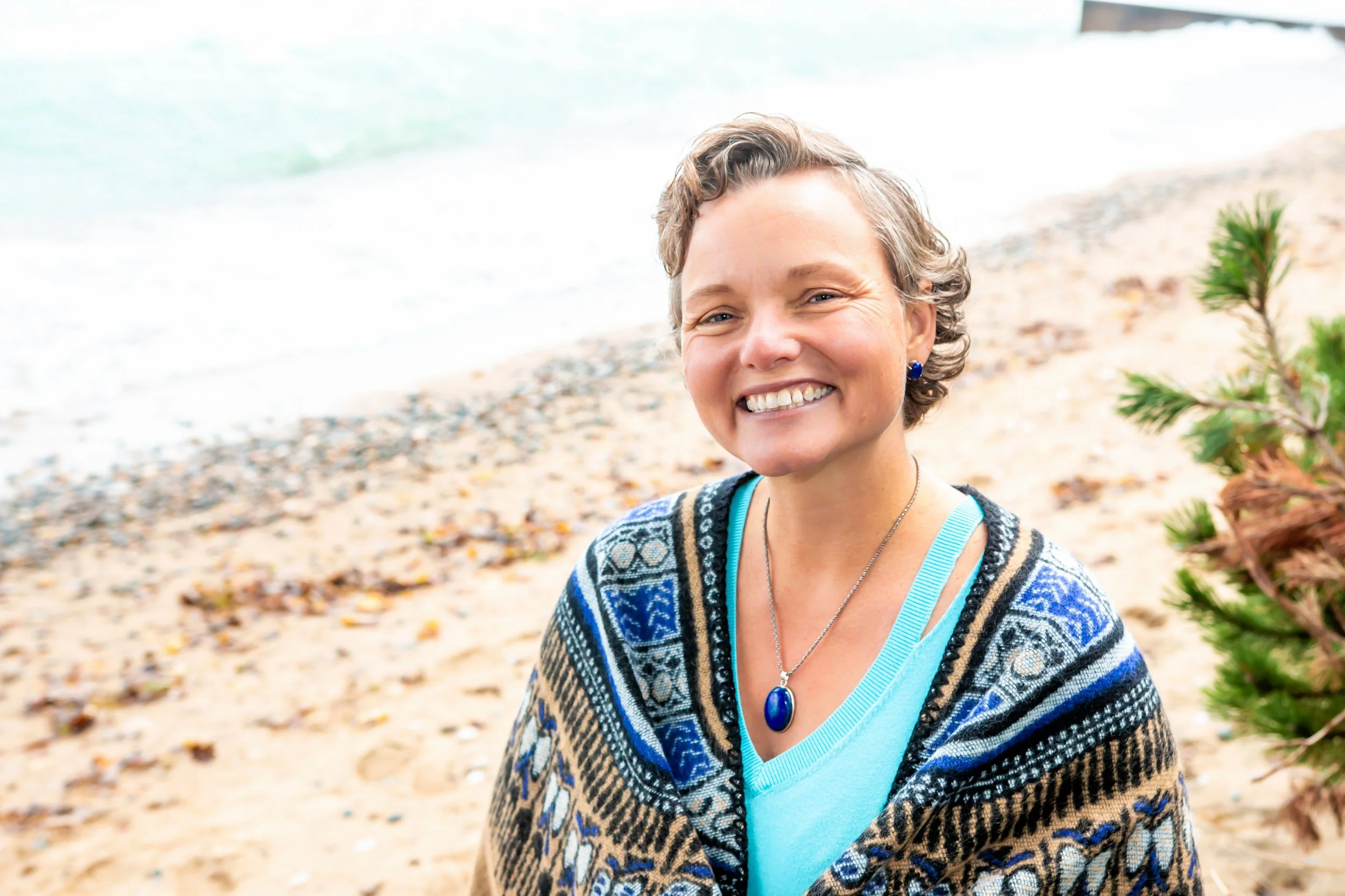 A woman with short curly hair smiling at the camera on a sandy beach, wearing a blue top, a patterned shawl, a necklace with a large blue pendant, and earrings, with sea and a small pine tree in the background.