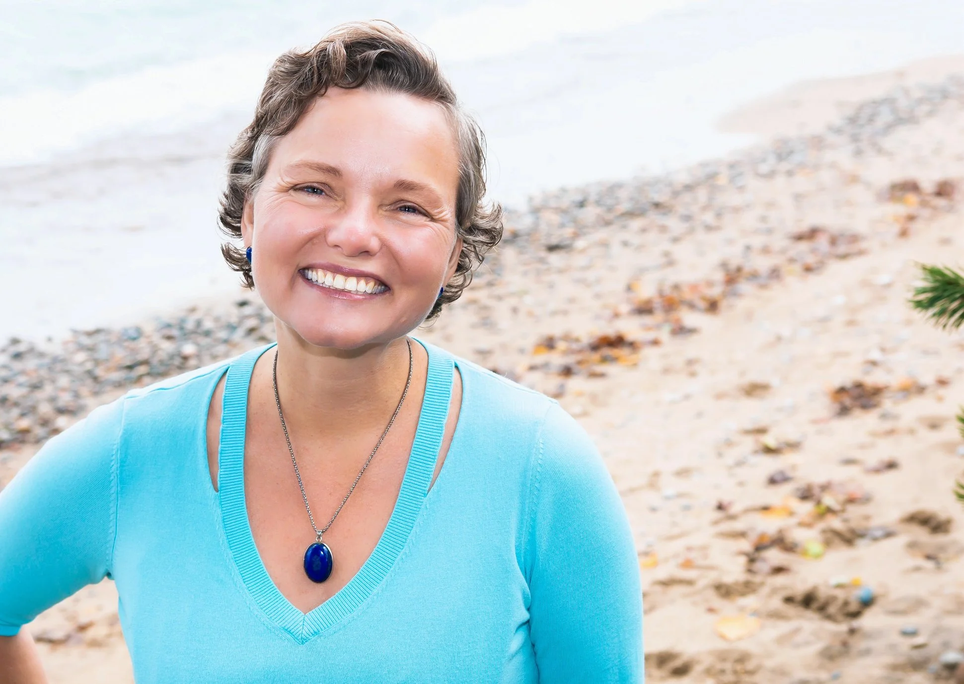 A woman with short, curly hair smiling at the camera on a beach, wearing a turquoise top and a matching necklace with a large oval blue pendant.