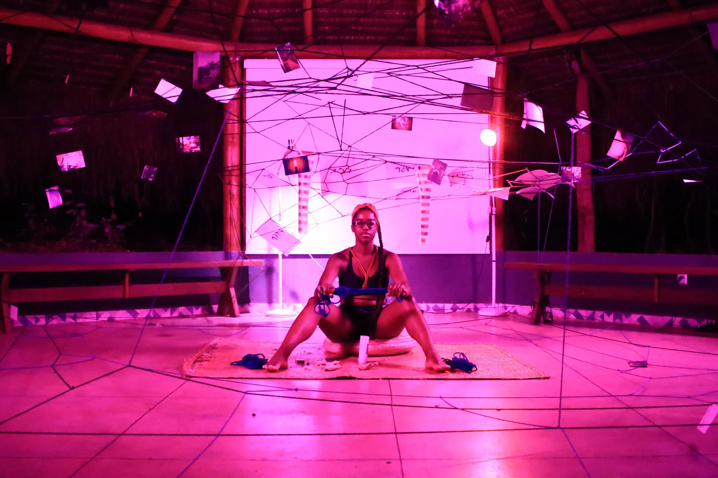 Une personne assise sur un tapis, tenant une corde, dans une pièce munie de filaments suspendus et de photos accrochées, éclairée en violet.