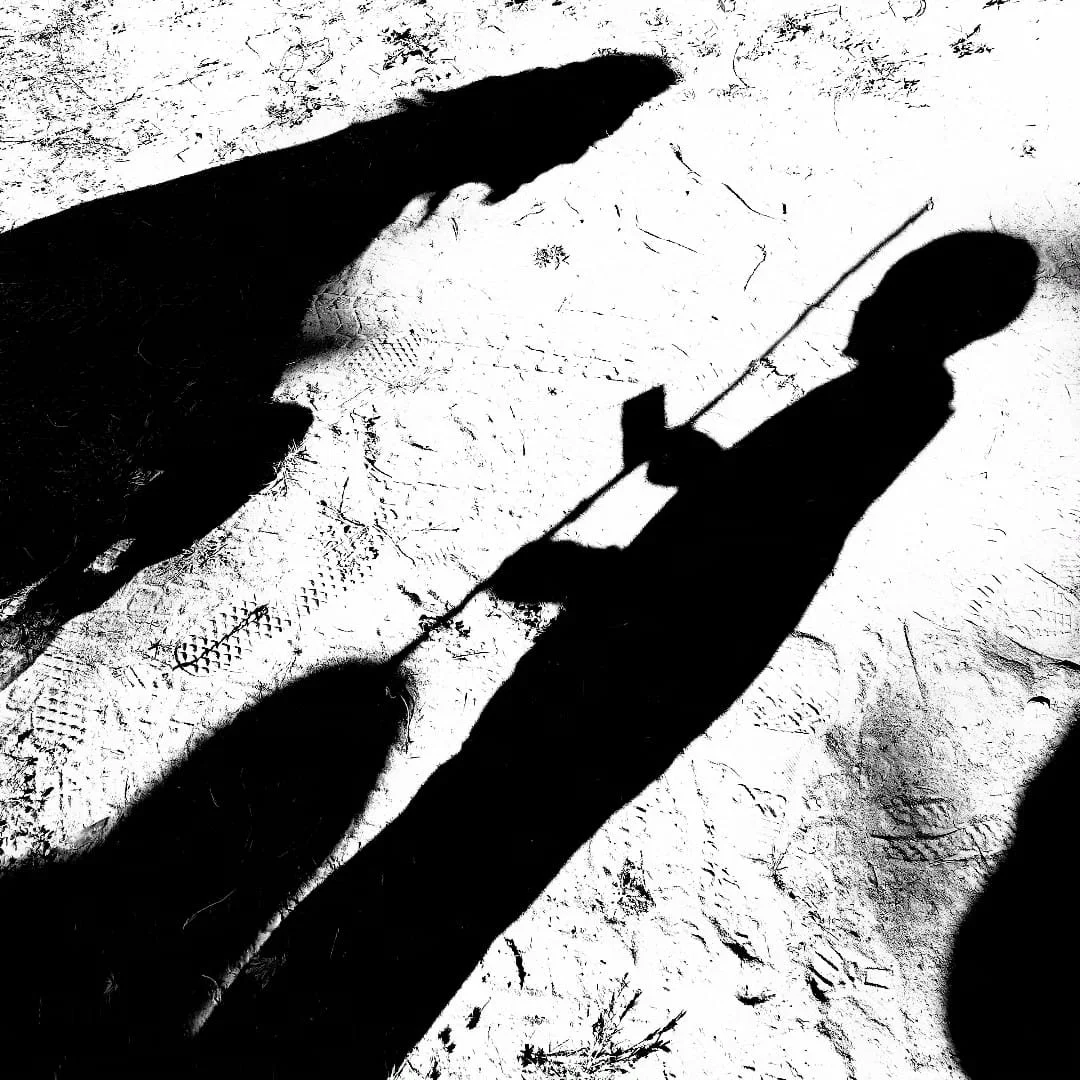 Ombres de deux personnes sur un sol sablonneux, une portant une boussole ou un bâton, la surface du sol est marquée par des empreintes et des traces de pas.