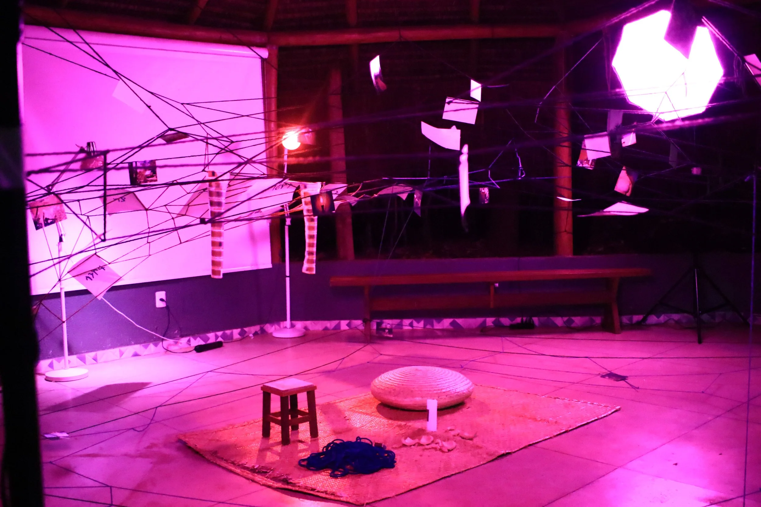 Une installation artistique avec des fils noirs tendus dans l'espace, éclairés en violet. Des photos suspendues et des bandes de papier sont accrochées aux fils. Le sol est recouvert d'un tapis avec un petit tabouret en bois, un coussin rond, une cor