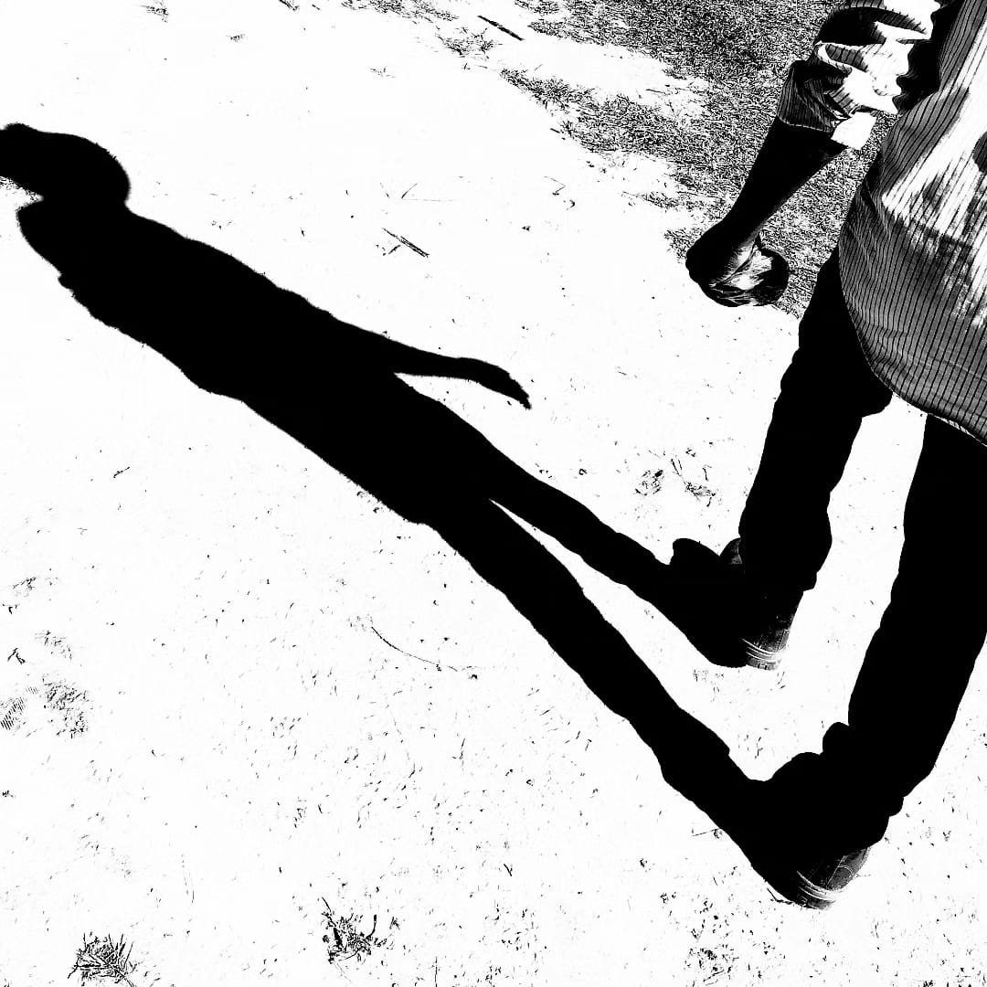 Une personne debout sur un sol sableux avec un long ombre projetée devant elle, vue de face. La personne porte un pantalon noir et une veste à rayures.