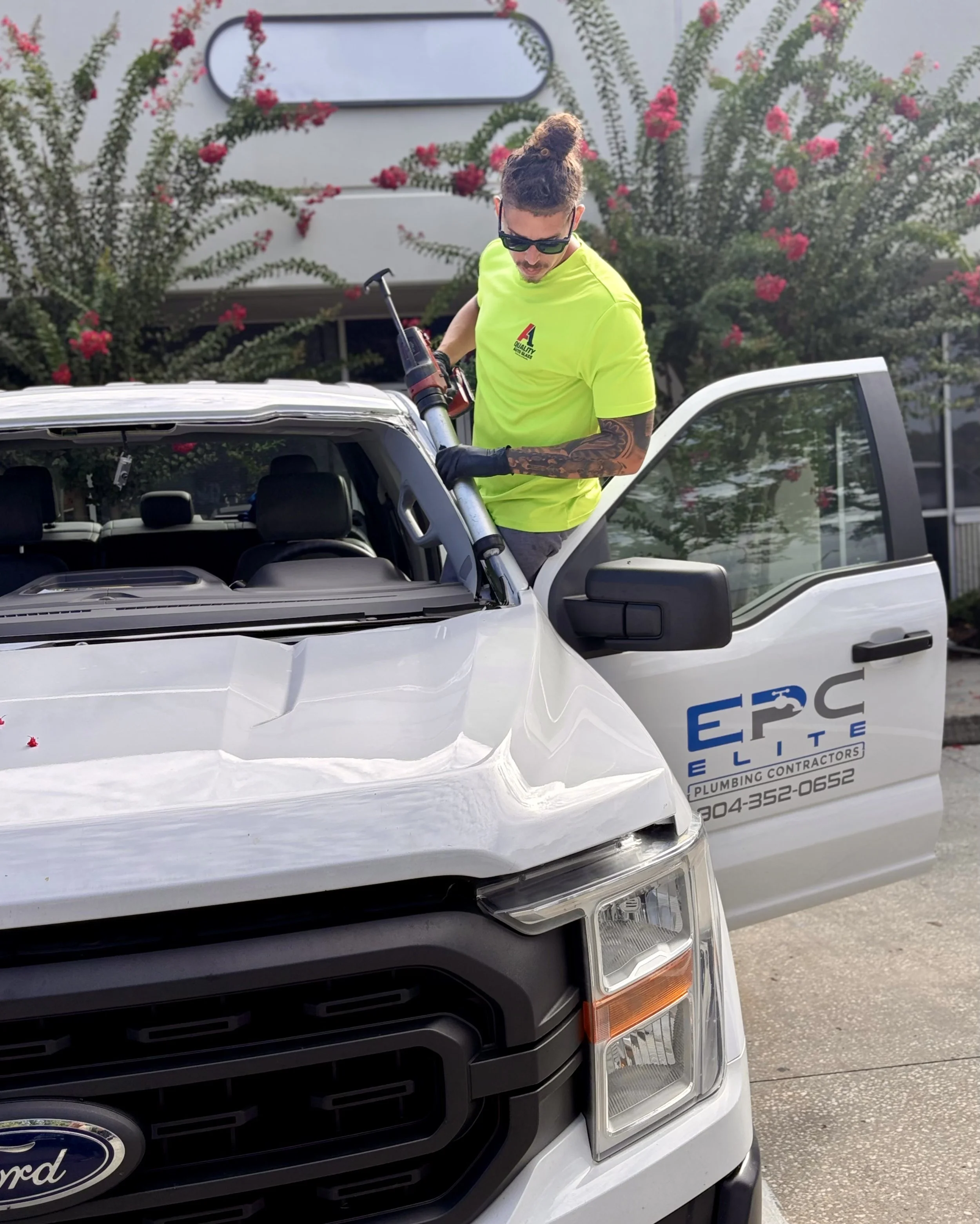 A man with tattoos and sunglasses wearing a bright yellow shirt, operating a hydraulic lift tool on the front of a white Ford pickup truck. The truck has contact information for a plumbing contractor named EPG Elite on the side, with a background of flowering bushes with pink and red flowers.