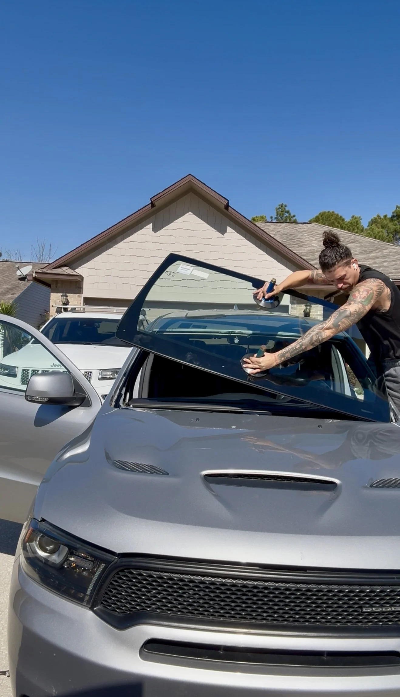 A woman with tattoos and a bun hairstyle cleaning the windshield of a silver Dodge car in a driveway under a clear blue sky.