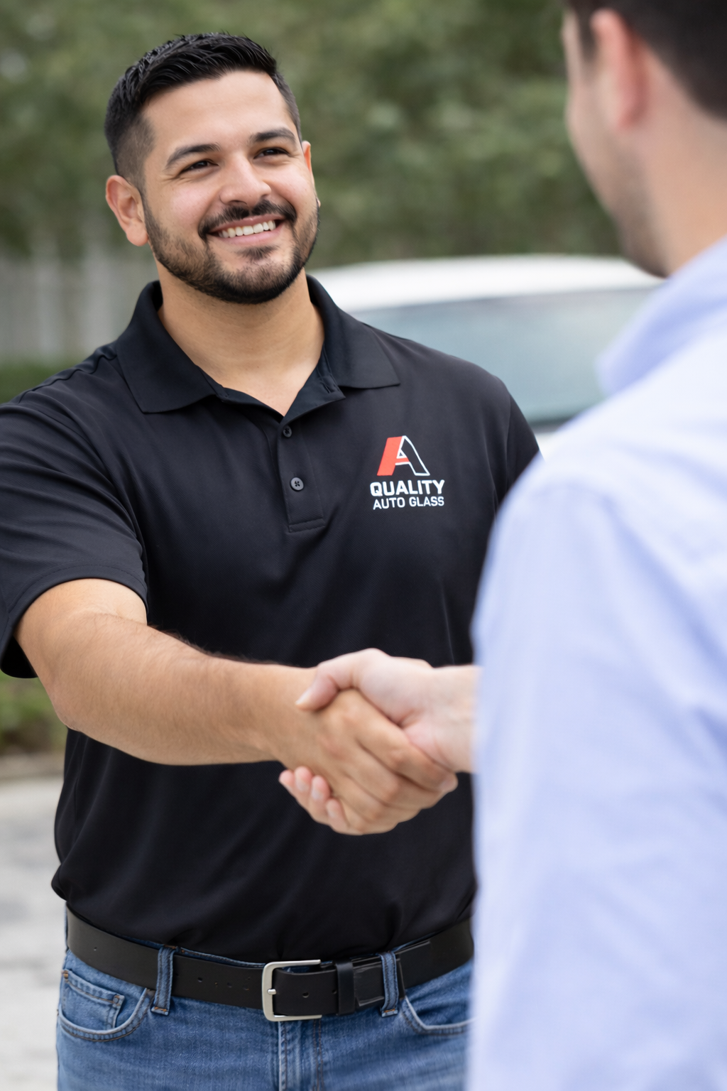 A man with dark hair and a beard smiling and shaking hands with a customer outdoors. The man is wearing a black polo shirt with a logo that reads 'Quality Auto Glass'.