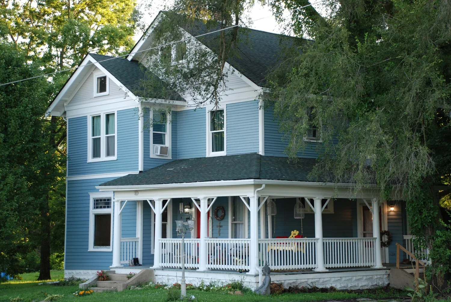 A two-story blue house with white trim, front porch, and a porch swing, surrounded by green trees and grass, with a small garden and decorative statues in the yard.