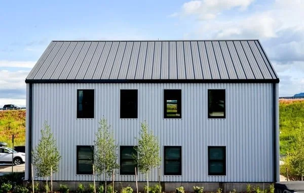 Modern two-story metal building with four windows on each floor, surrounded by small trees and a field under a partly cloudy sky.