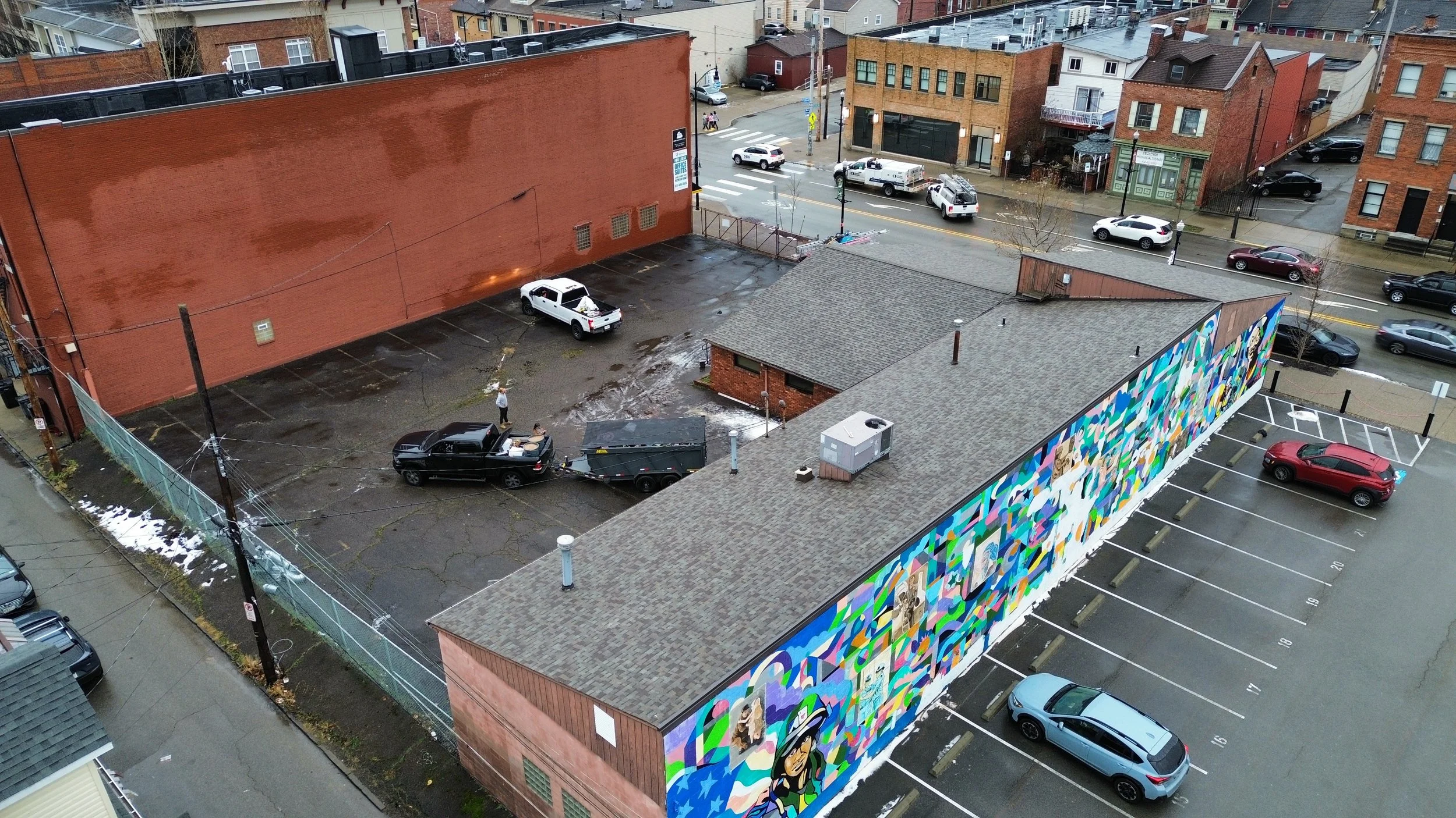 Aerial view of a city alley with parked cars, a mural on the building wall, and pedestrians walking on the street.
