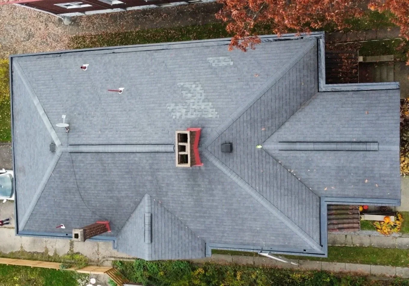 Aerial view of a house's roof with multiple vents, chimneys, and a satellite dish, surrounded by trees and a sidewalk.