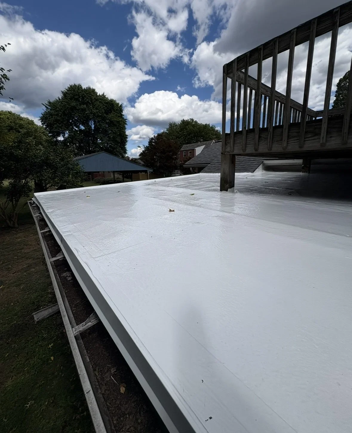 Roof coated with white reflective sealant or paint, with a wooden deck railing visible on the right, and a blue sky with clouds and trees in the background.