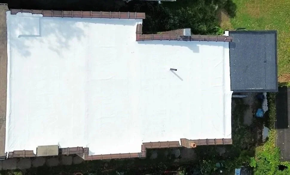 Aerial view of a building roof with a white reflective material, brick chimneys on the corners, and surrounding greenery.