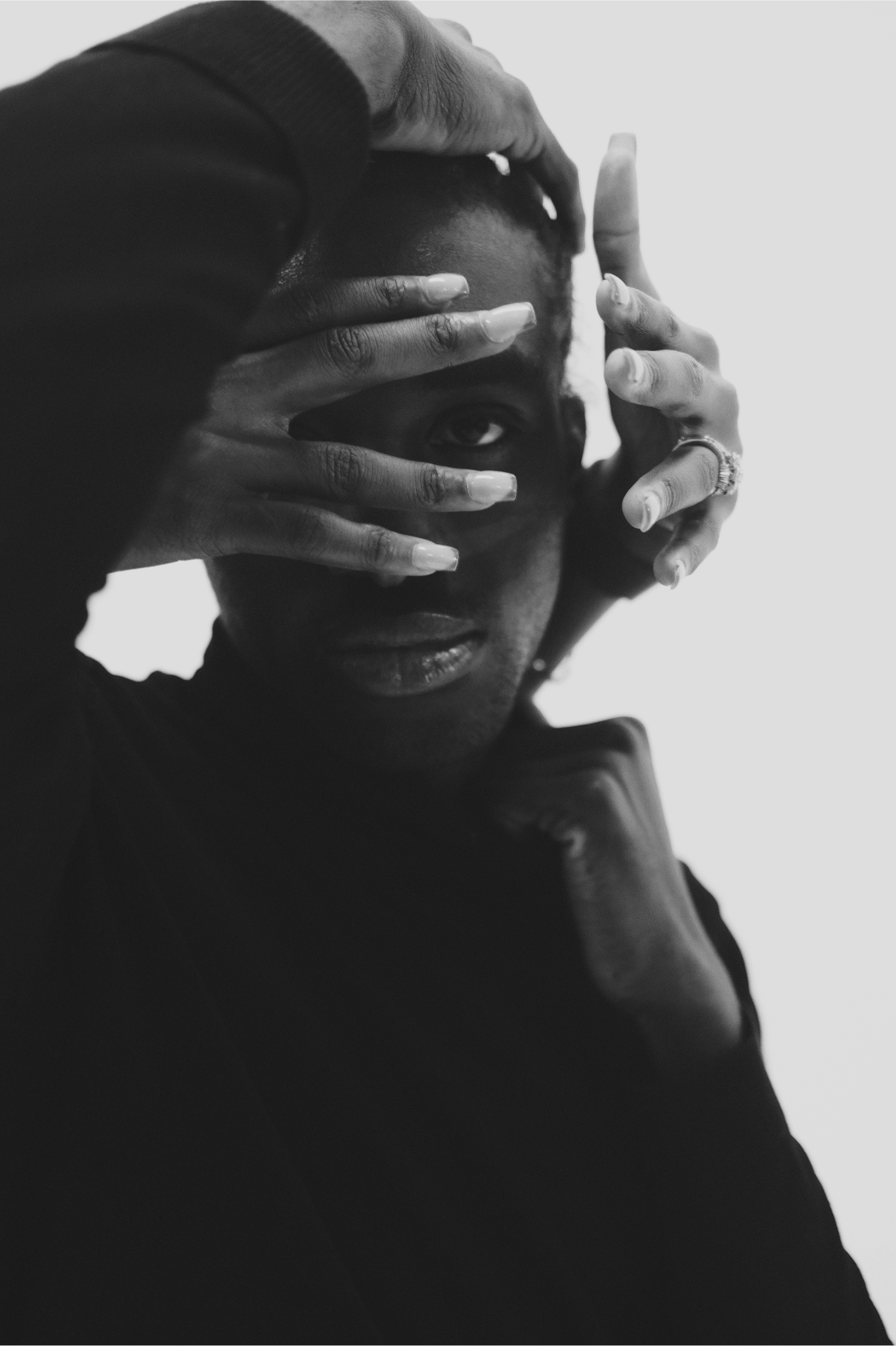 A black and white portrait of a person with short hair, partially covering their face with their hands, looking directly at the camera.